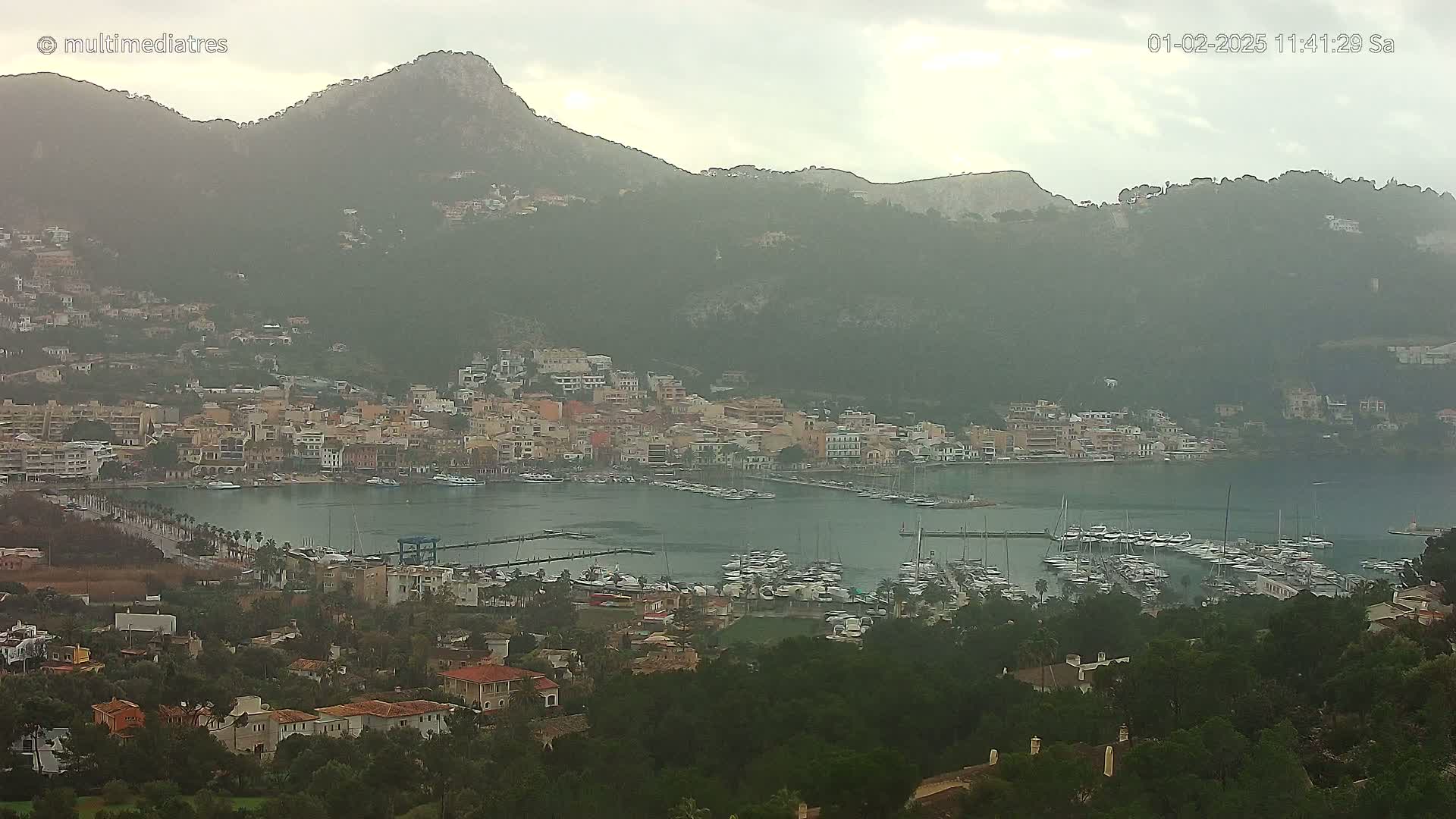 A hazy, overcast day reveals a coastal town nestled in a harbor, surrounded by lush green hills and mountains.