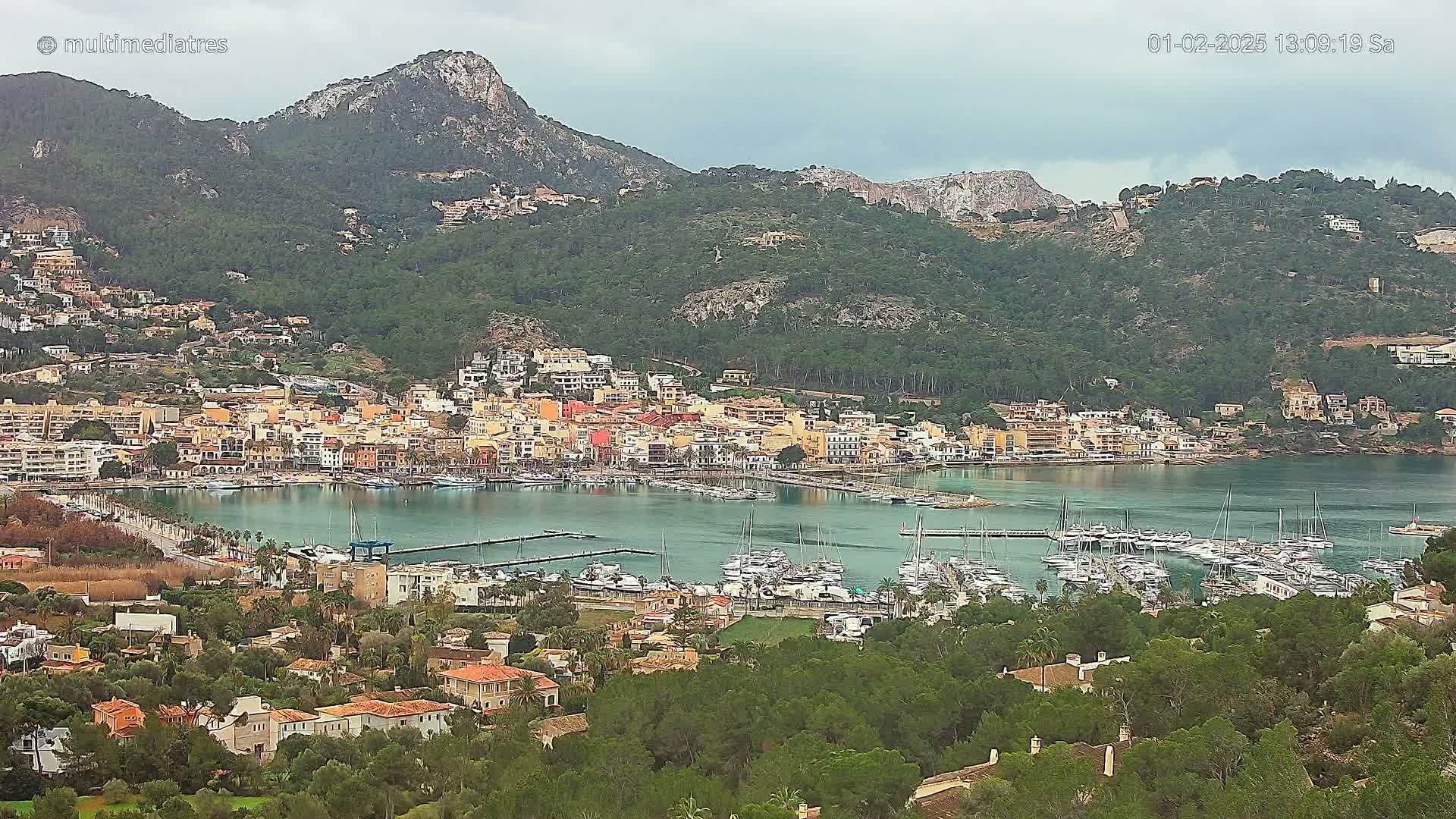A coastal town with a marina full of sailboats nestled at the base of a mountain range under an overcast sky.