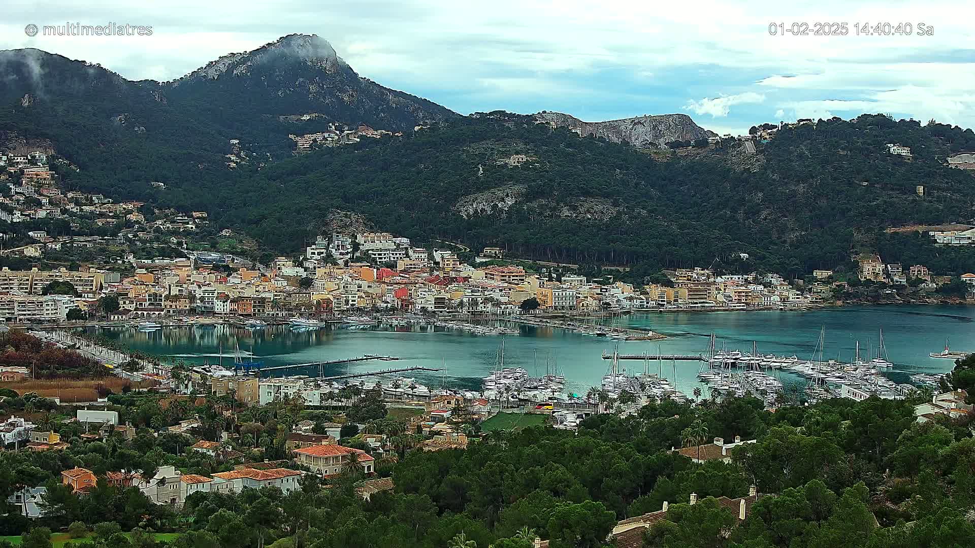 A coastal town with a marina full of boats sits nestled at the base of a mountain range under a partly cloudy sky.