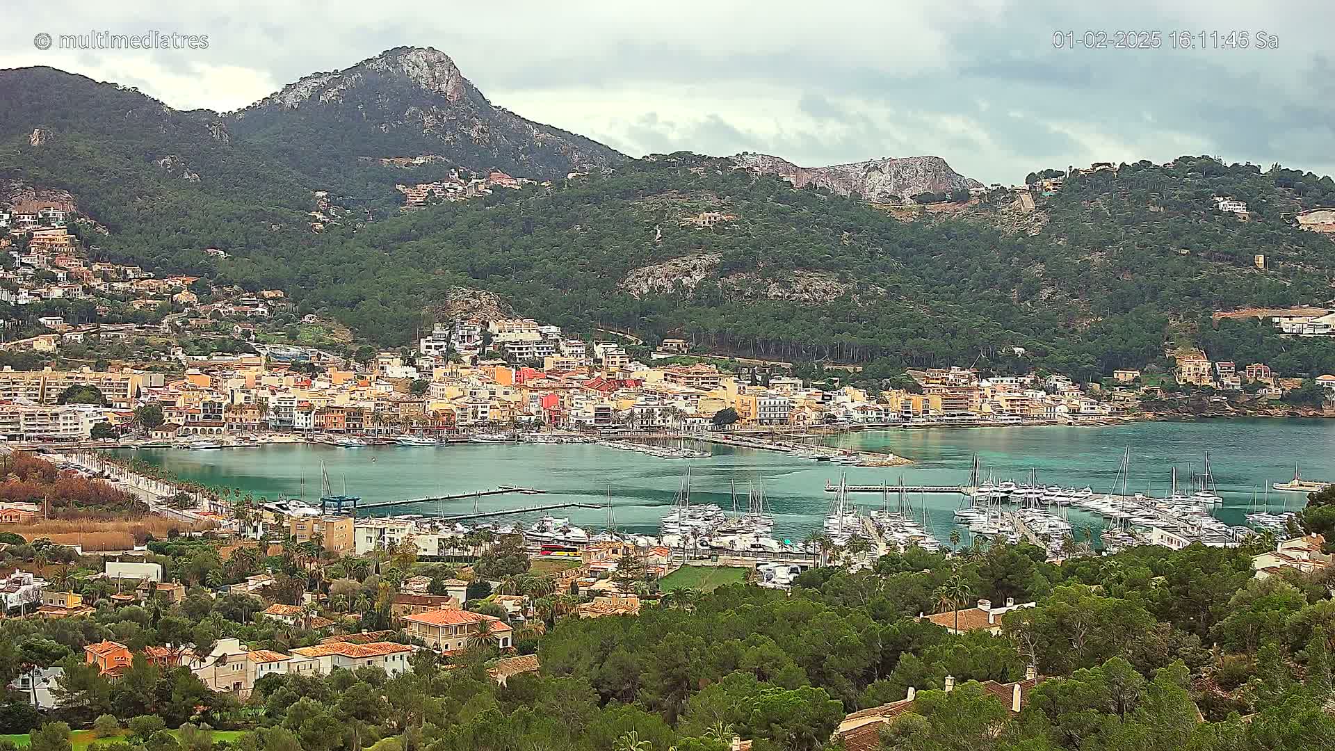 A coastal town nestled between a mountain range and a calm, green-blue bay filled with sailboats under an overcast sky.