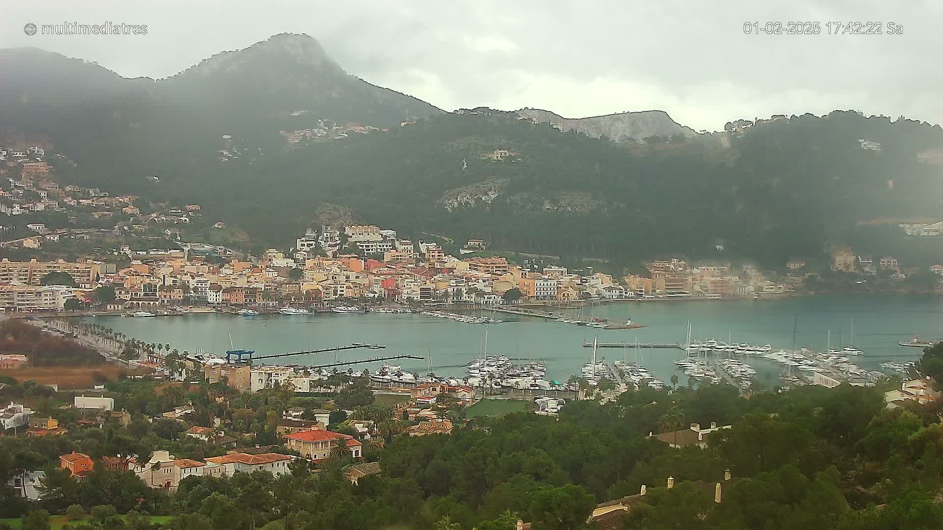 A coastal town nestled between mountains, with a marina full of boats, is seen on an overcast day.