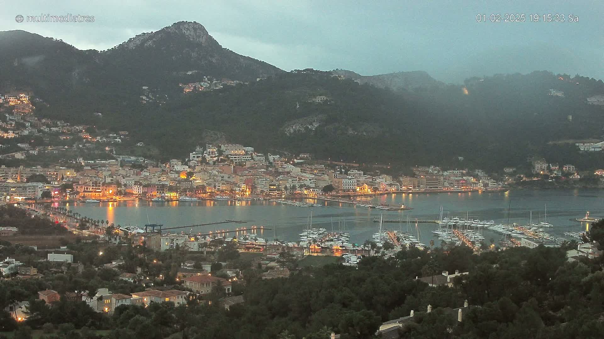 A nighttime view of a harbor town nestled between hills under a cloudy sky, with many boats moored in the harbor and lights illuminating the buildings.