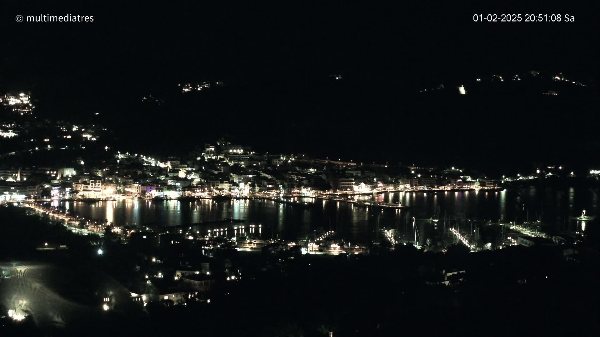 A nighttime, high-angle view shows a coastal town brightly lit along a calm body of water, with boats and a marina visible.