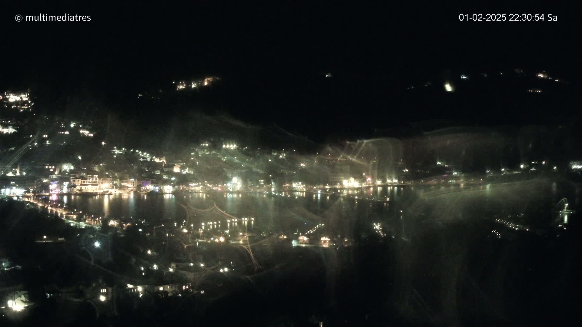 A nighttime, blurry view of a town situated beside a body of water, with its lights reflecting on the water's surface.