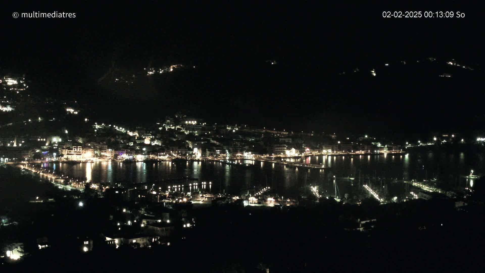 A nighttime, aerial view shows a coastal town illuminated with lights, nestled along a dark body of water, with boats and docks visible.