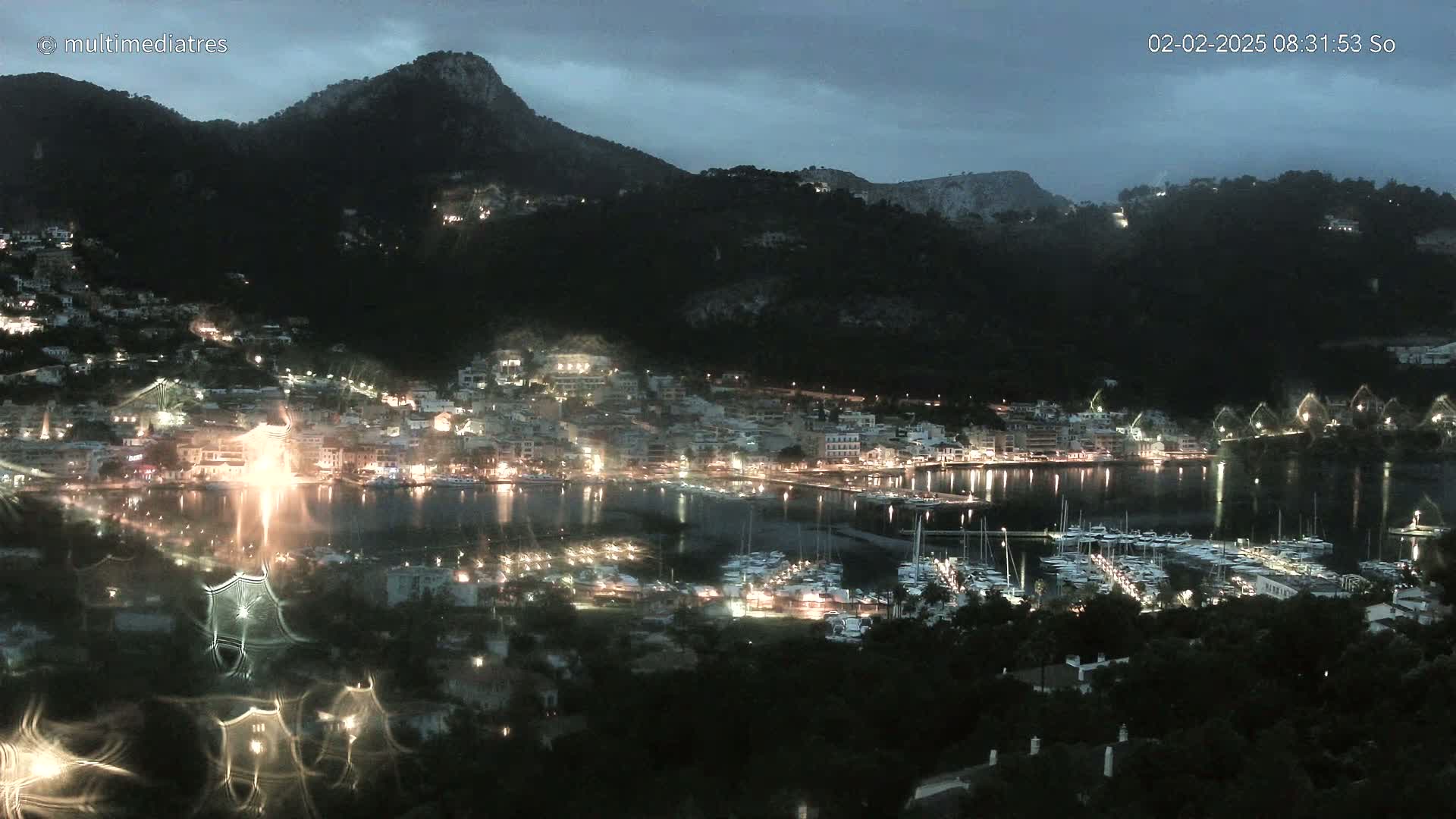 A nighttime view of a harbor town nestled between dark hills under an overcast sky, with many boats in the marina.