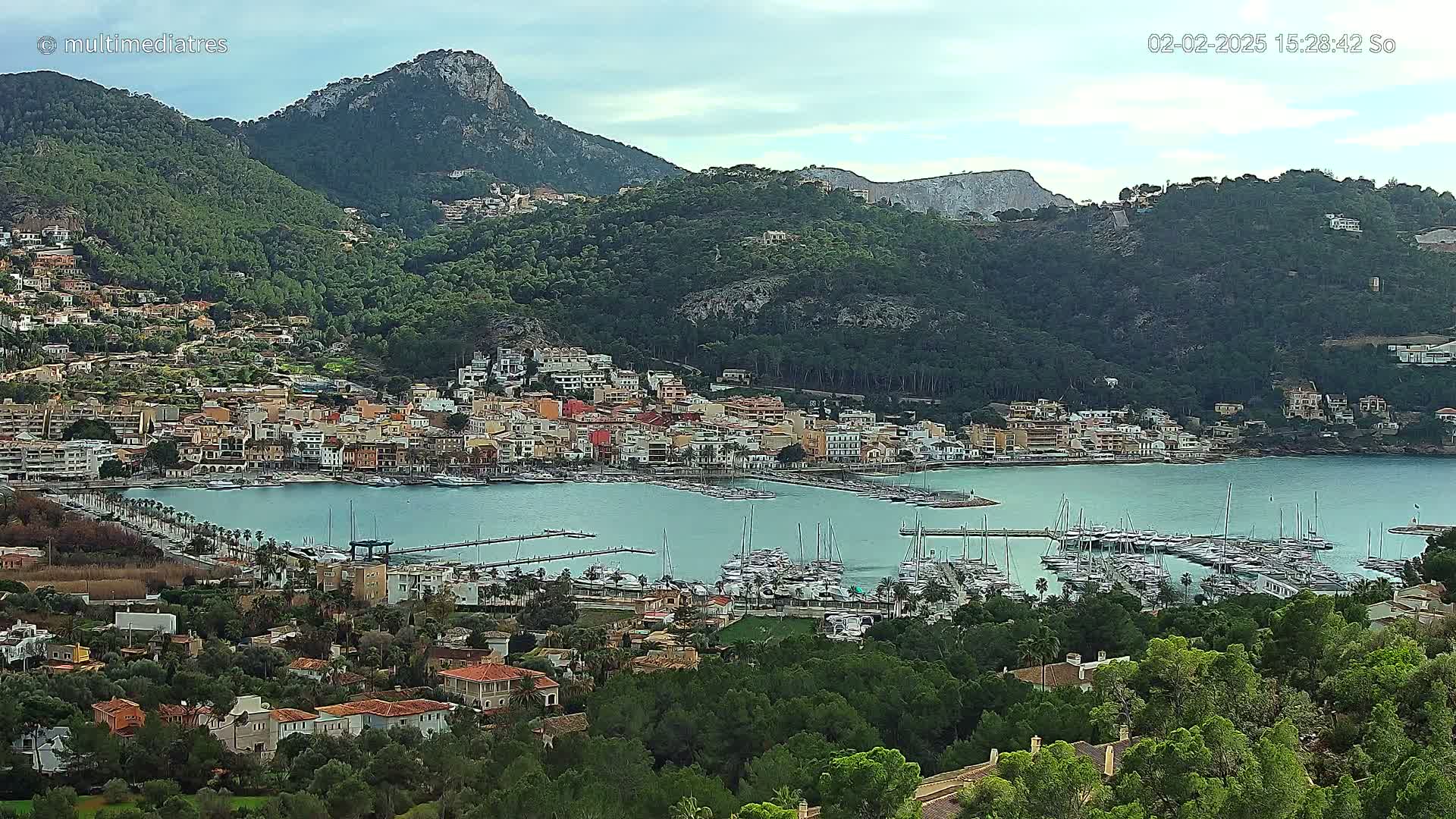A coastal town nestled in a verdant valley, bordered by mountains under a mostly cloudy sky, features a bustling marina filled with numerous boats.