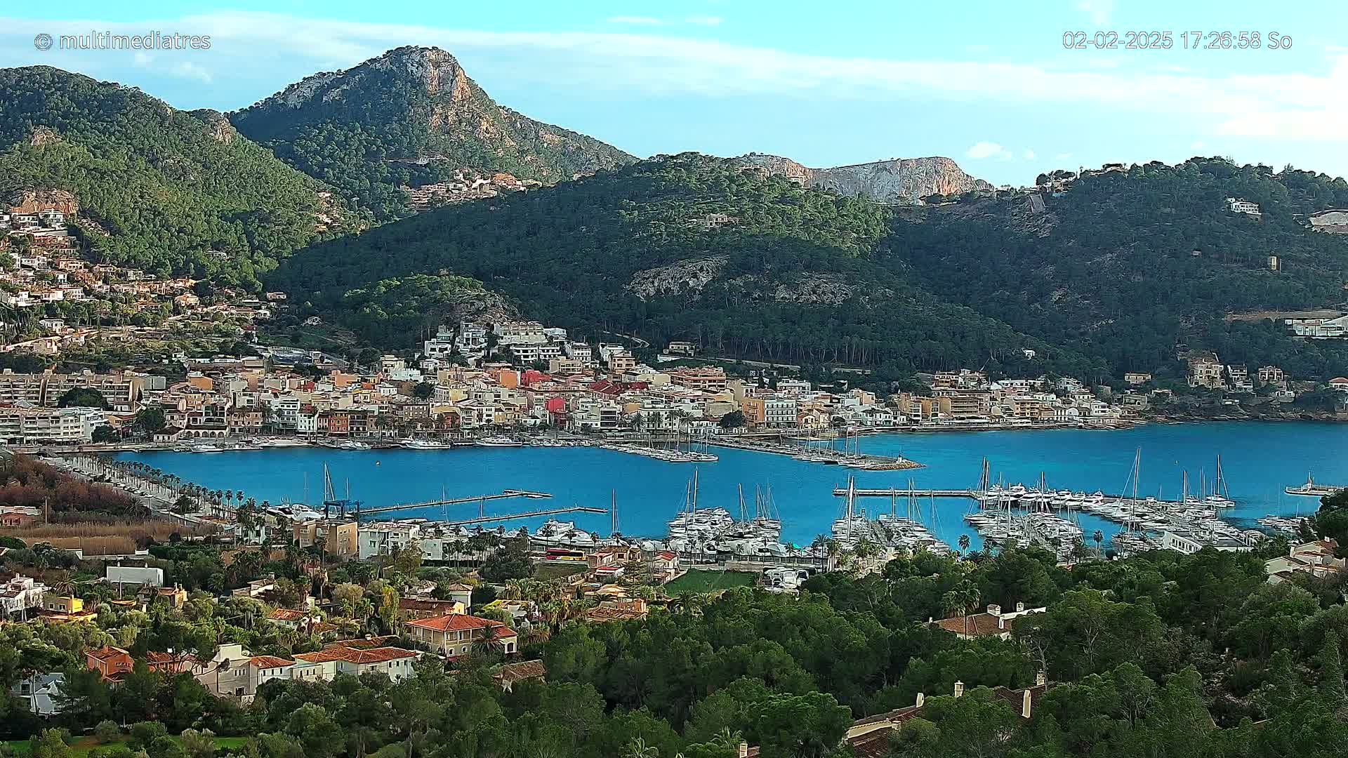 A picturesque coastal town nestled between lush green hills, featuring a vibrant marina filled with numerous boats under a clear, sunny sky.
