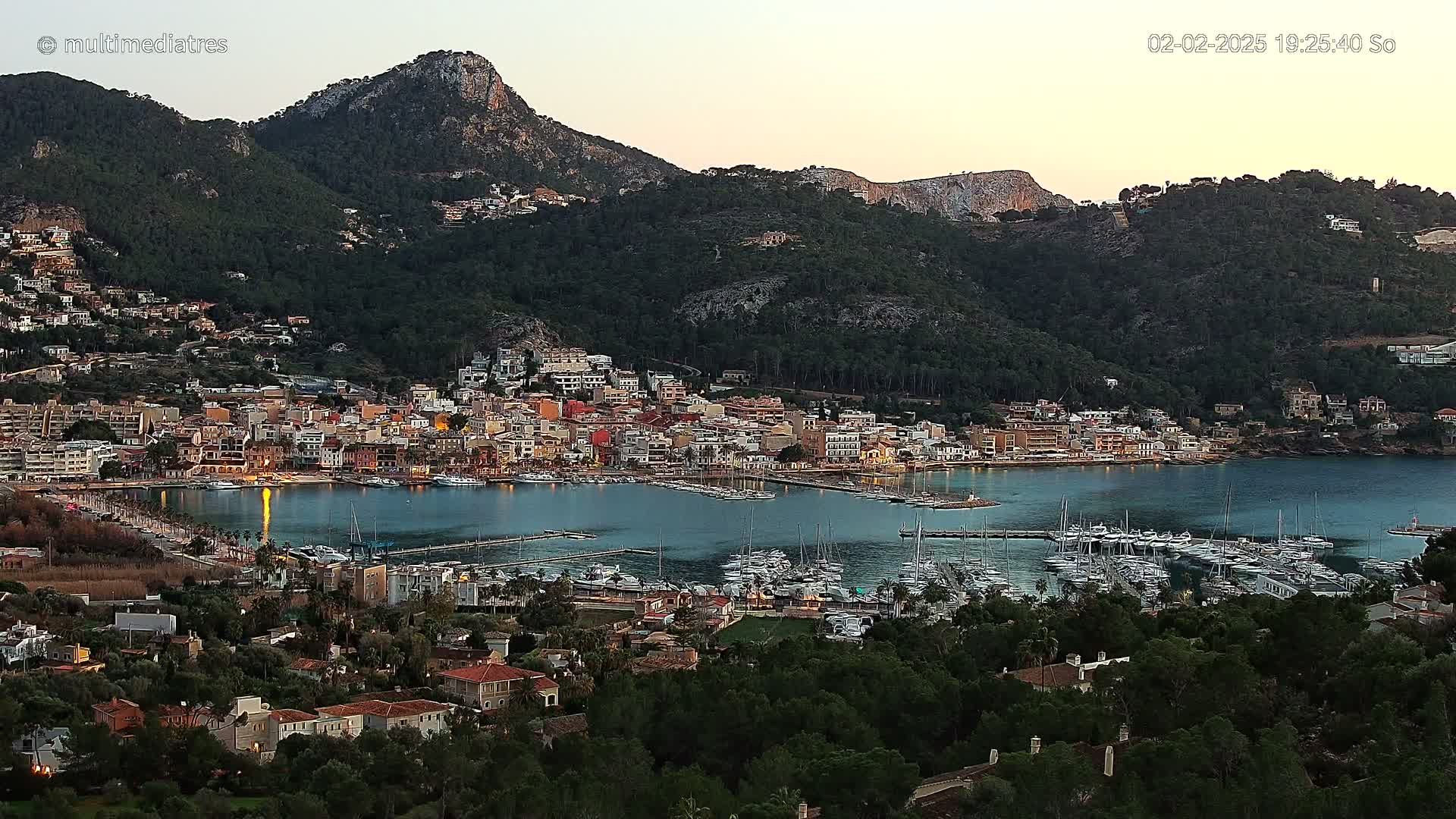 A coastal town nestled between a calm bay filled with sailboats and dark green hills under a twilight sky.