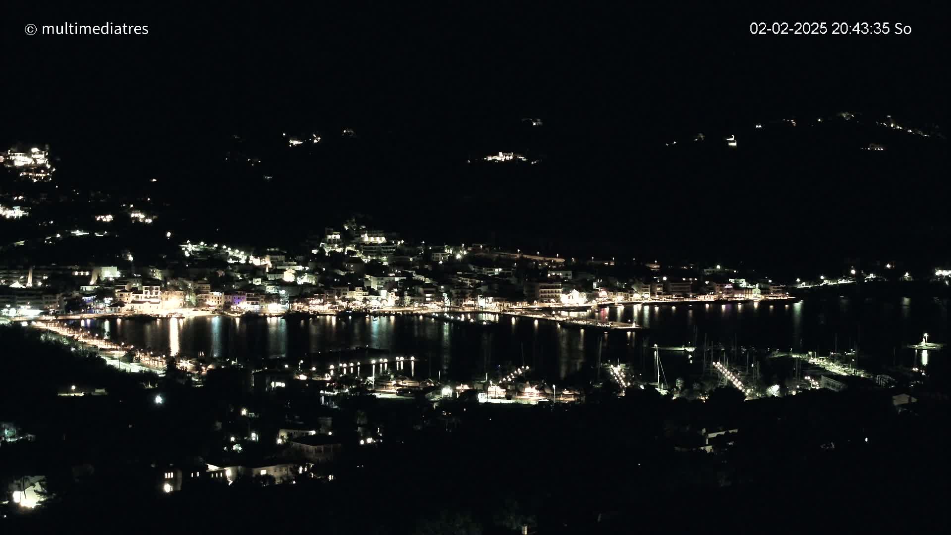 A nighttime, aerial view shows a coastal town illuminated with lights surrounding a dark body of water filled with boats.