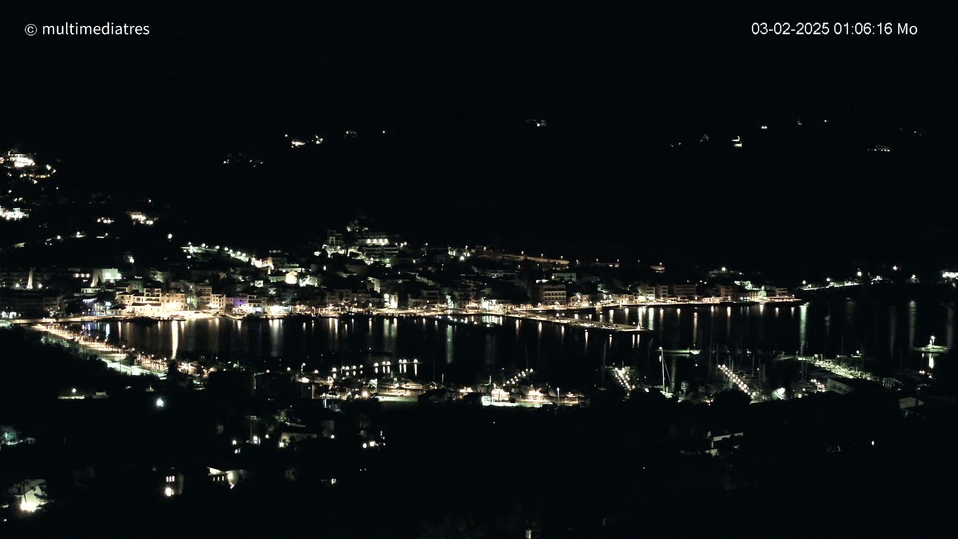 A nighttime view of a town's harbor and waterfront, illuminated by lights reflecting on calm water.