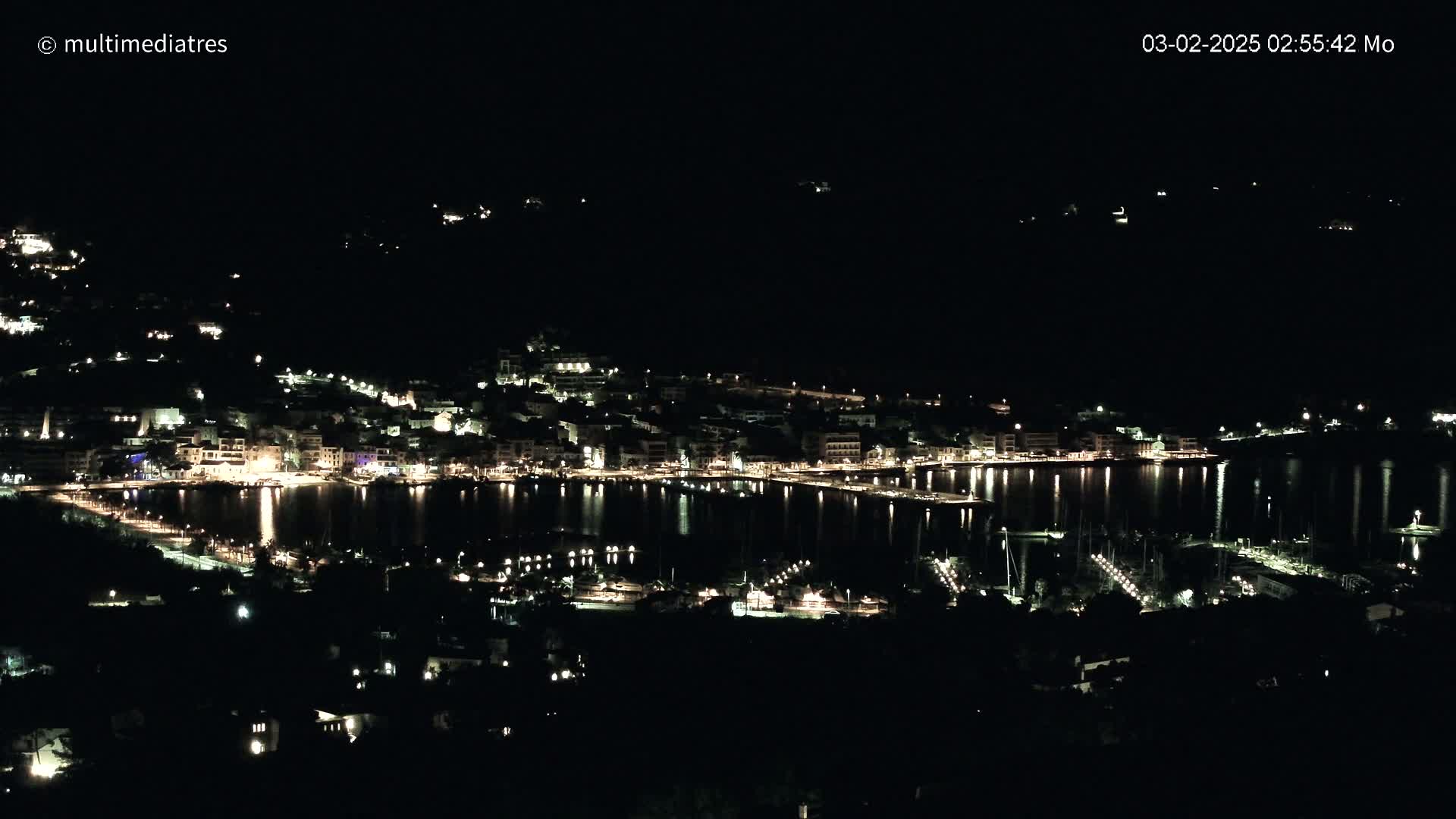 A nighttime view of a harbor town, brightly lit, with calm water reflecting the lights.