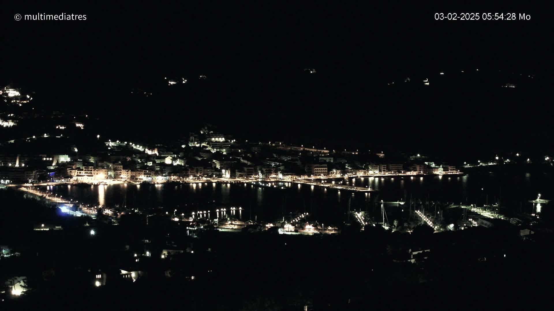 A nighttime aerial view shows a coastal town with its lights illuminating the harbor and surrounding buildings under a clear, dark sky.