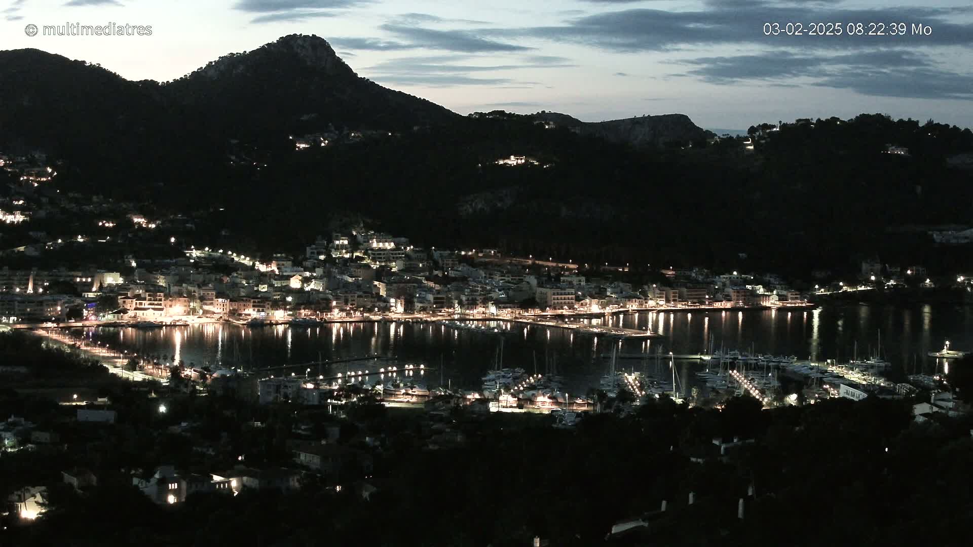 A nighttime view of a harbor town nestled at the base of a dark mountain under a twilight sky.