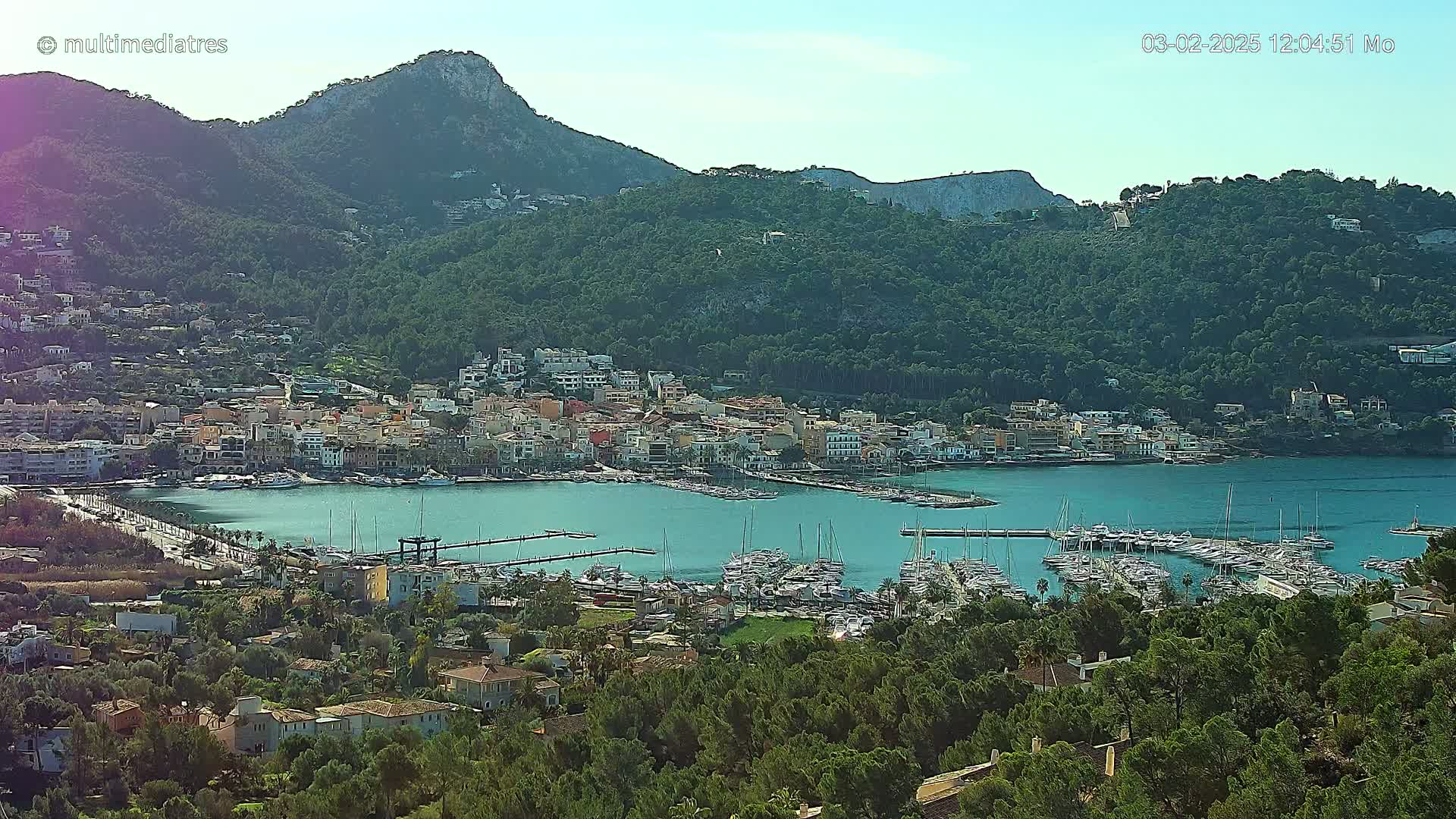 A coastal town nestled between green hills and a calm, blue bay filled with numerous boats under a clear sky.