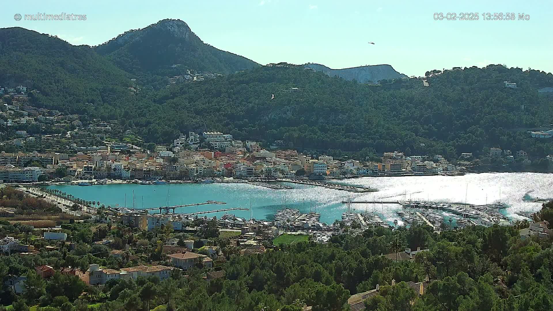 A picturesque coastal town nestled in a mountainous region with a marina full of boats, under a clear, sunny sky.