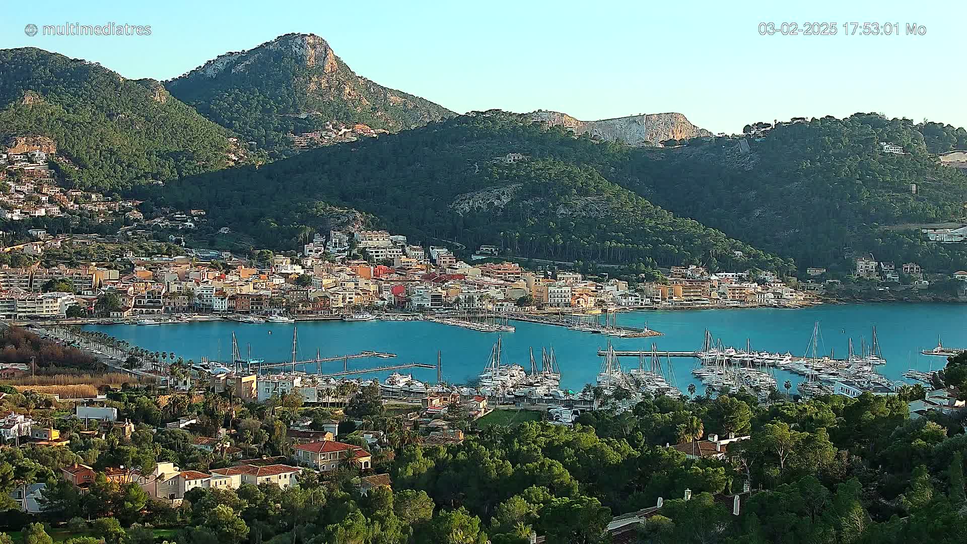 A coastal town nestled between verdant hills and a calm blue bay filled with sailboats, under a clear sky.