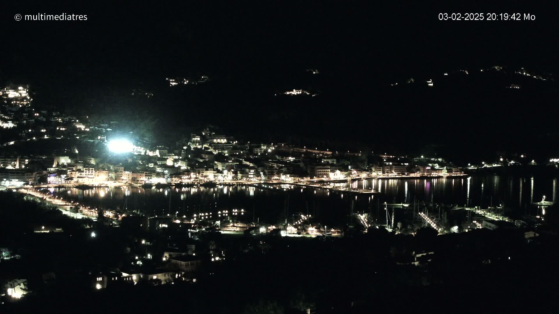 A nighttime, high-angle view of a coastal town illuminated by lights, reflecting on the calm water of a harbor filled with boats.