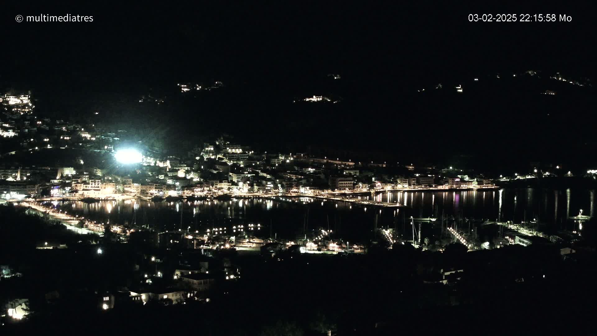 A nighttime view of a coastal town illuminated by lights, reflecting on a calm body of water, with boats moored in a marina.