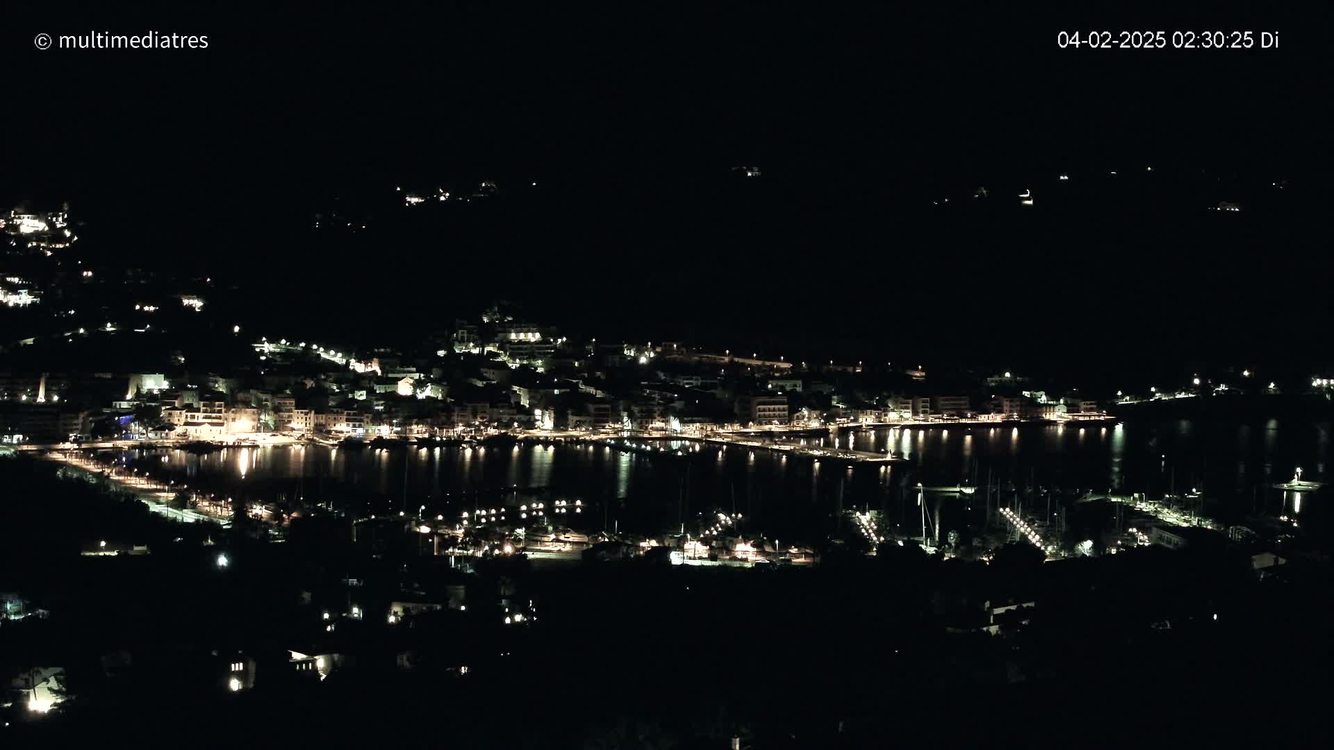 A coastal town at night, illuminated by many lights, is seen from a high vantage point under a dark, clear sky.