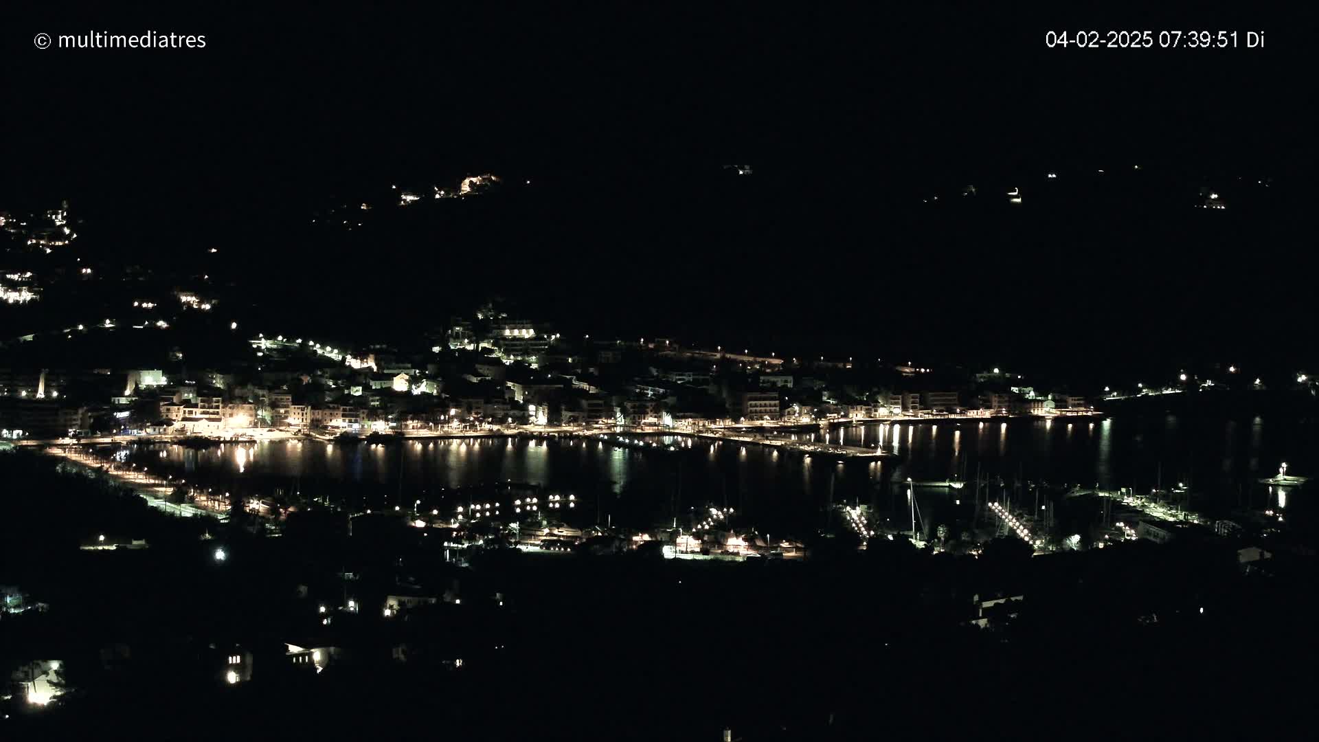 A nighttime view of a coastal town illuminated by lights, with a calm body of water reflecting the lights.