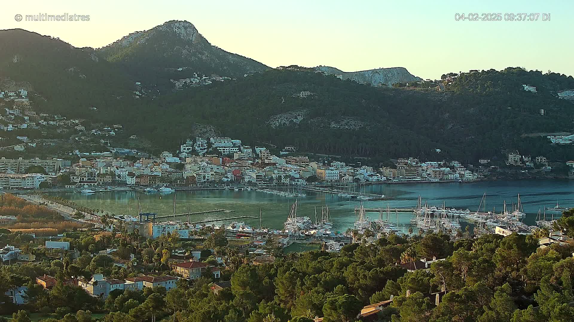 A coastal town nestled in a mountainous area with a calm harbor filled with numerous boats, under a clear, bright sky.