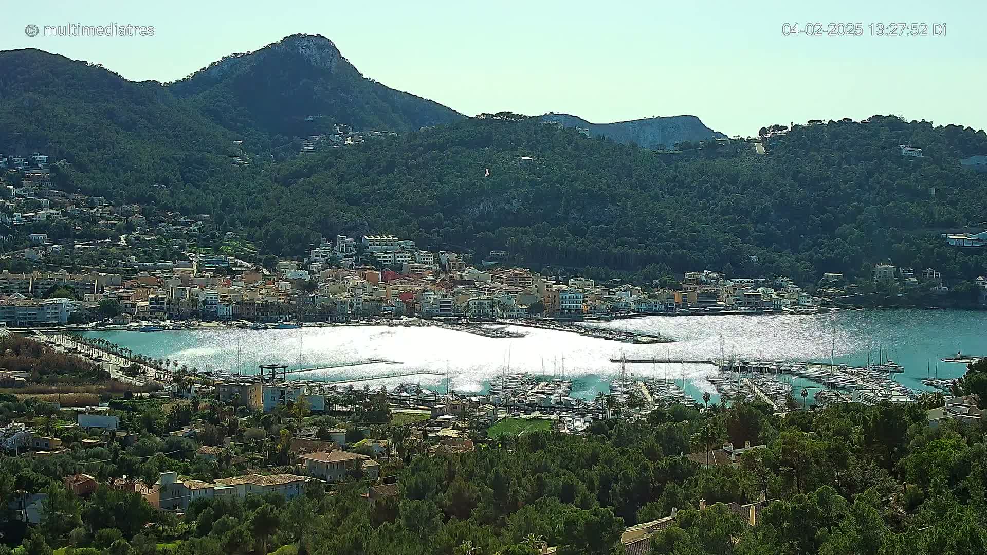 A coastal town nestled between green hills and a harbor filled with boats under a sunny sky.