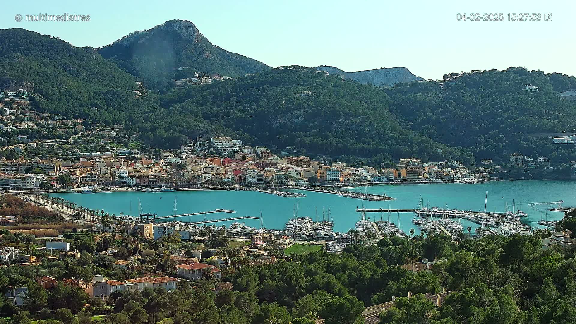A coastal town with a marina full of sailboats is nestled at the base of a large, green mountain under a clear sky.