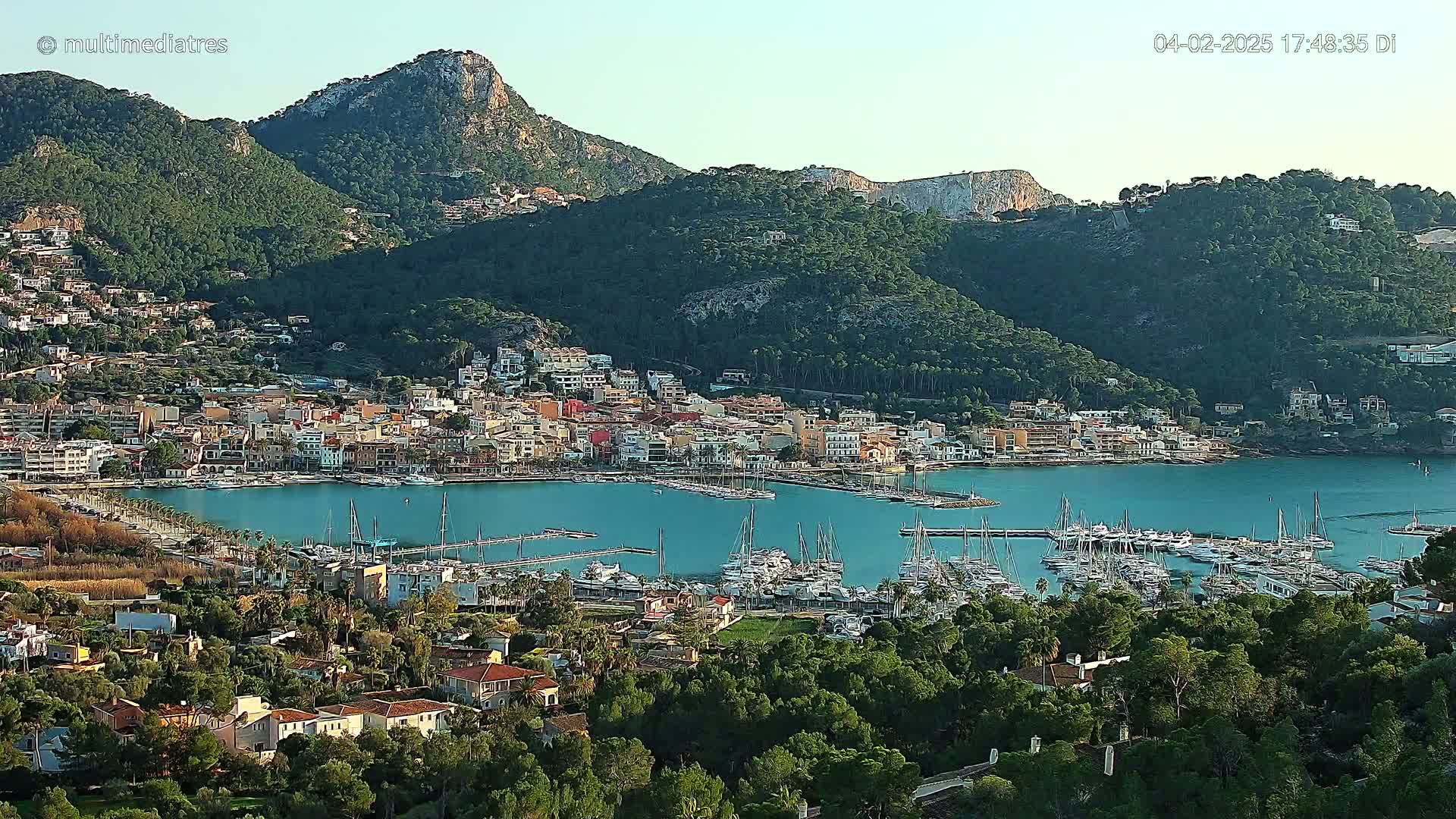 A picturesque coastal town nestled between verdant hills and a calm, blue harbor filled with sailboats, under a clear sky.