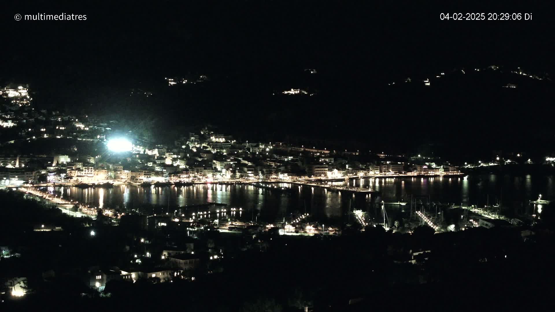 A nighttime, aerial view shows a harbor town with its lights reflected in the dark water, nestled against a dark hillside under a clear, night sky.