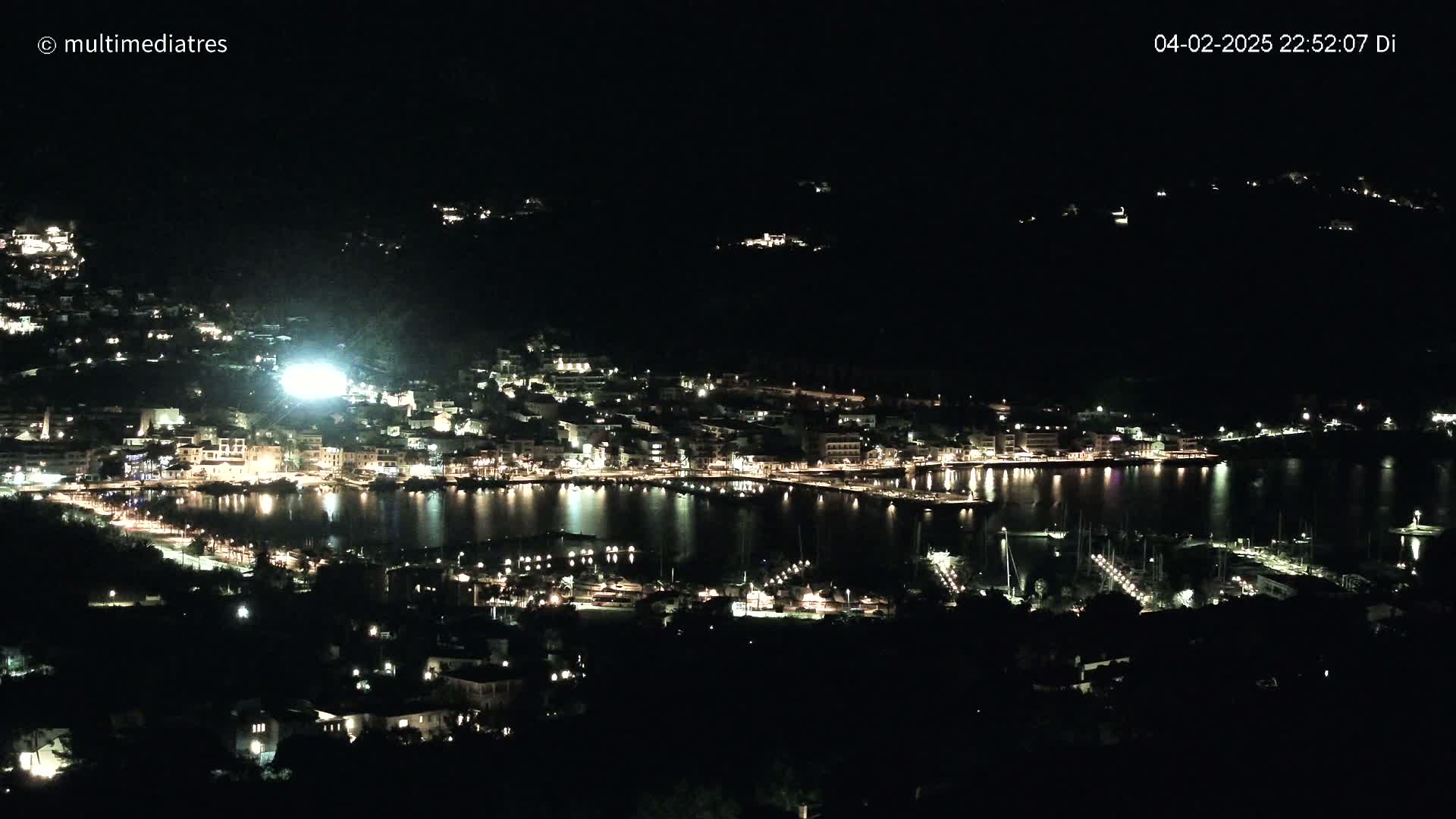 A nighttime view of a coastal town brightly lit, with its harbor filled with boats, under a clear, dark sky.