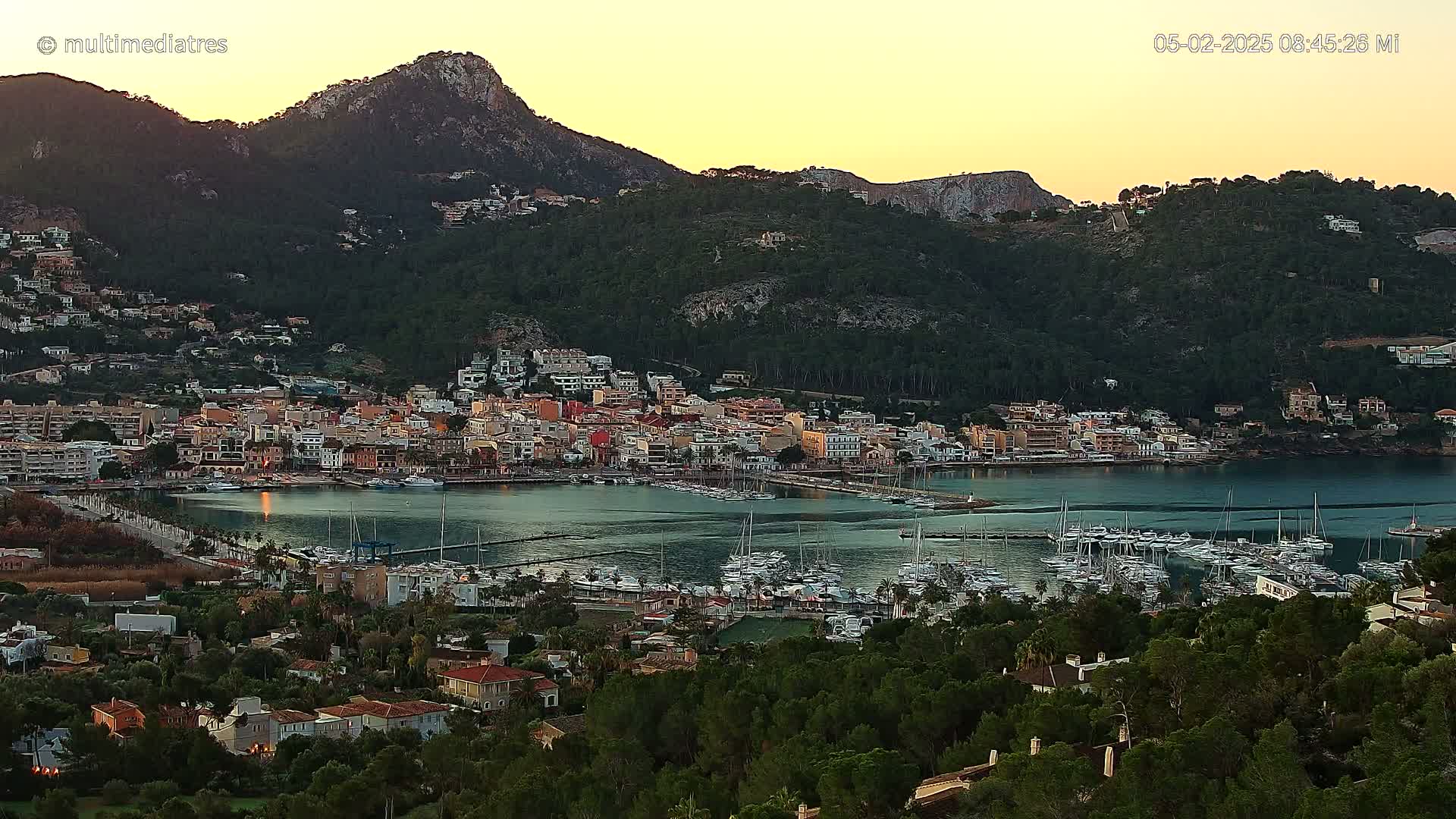 A coastal town nestled in a verdant hillside, with a calm harbor filled with sailboats, is bathed in the warm glow of a sunset.