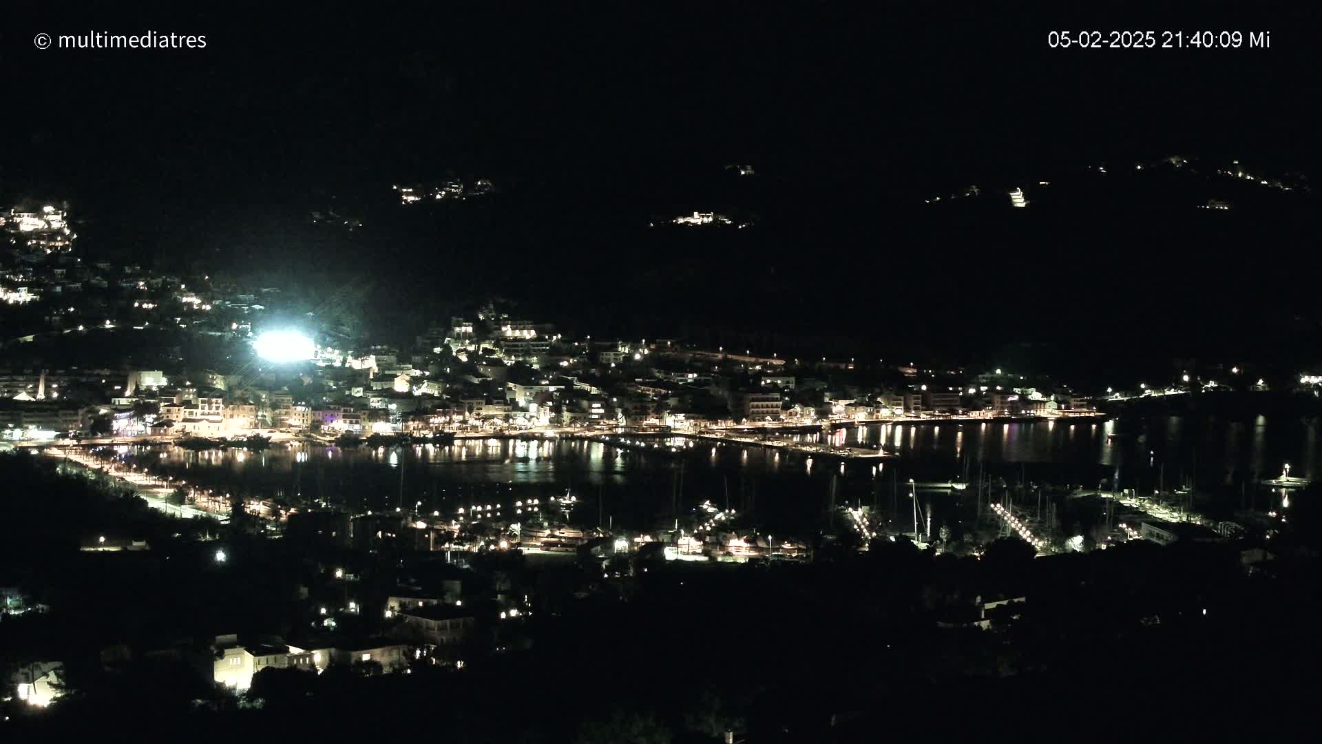 A nighttime aerial view of a coastal town with a harbor filled with boats, illuminated by many lights.