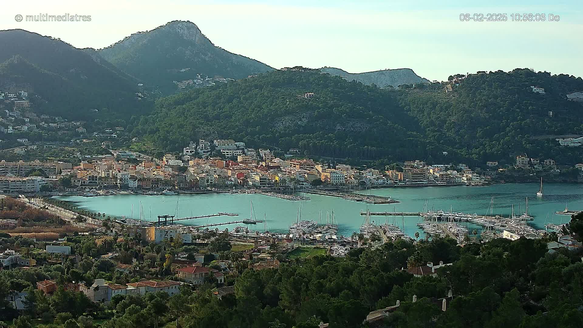 A picturesque coastal town nestled between green hills and a calm, turquoise harbor filled with numerous sailboats under a clear sky.