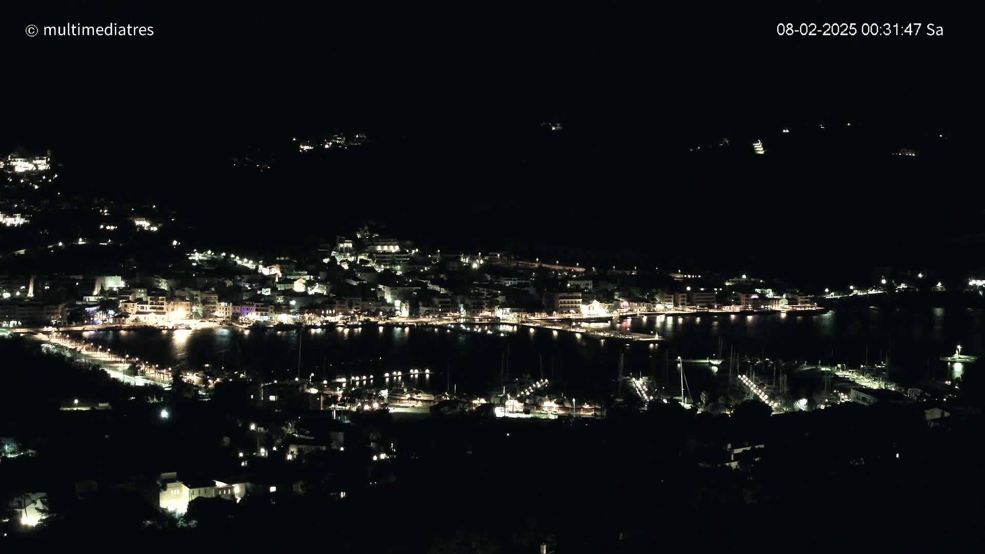 A coastal town at night, illuminated by many lights, is shown overlooking a harbor filled with boats.