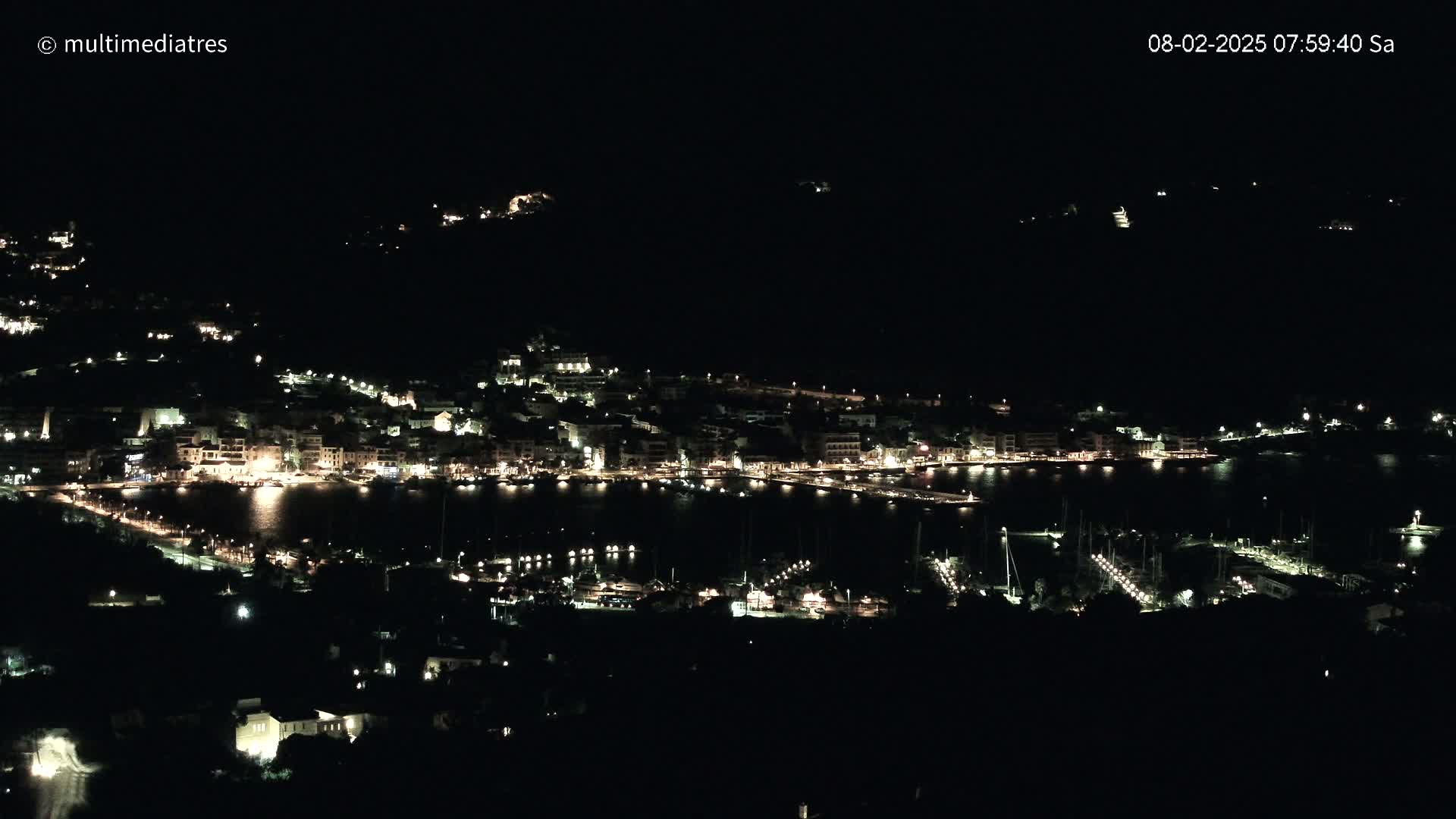 A coastal town at night is illuminated by lights from buildings and boats in a dark harbor.