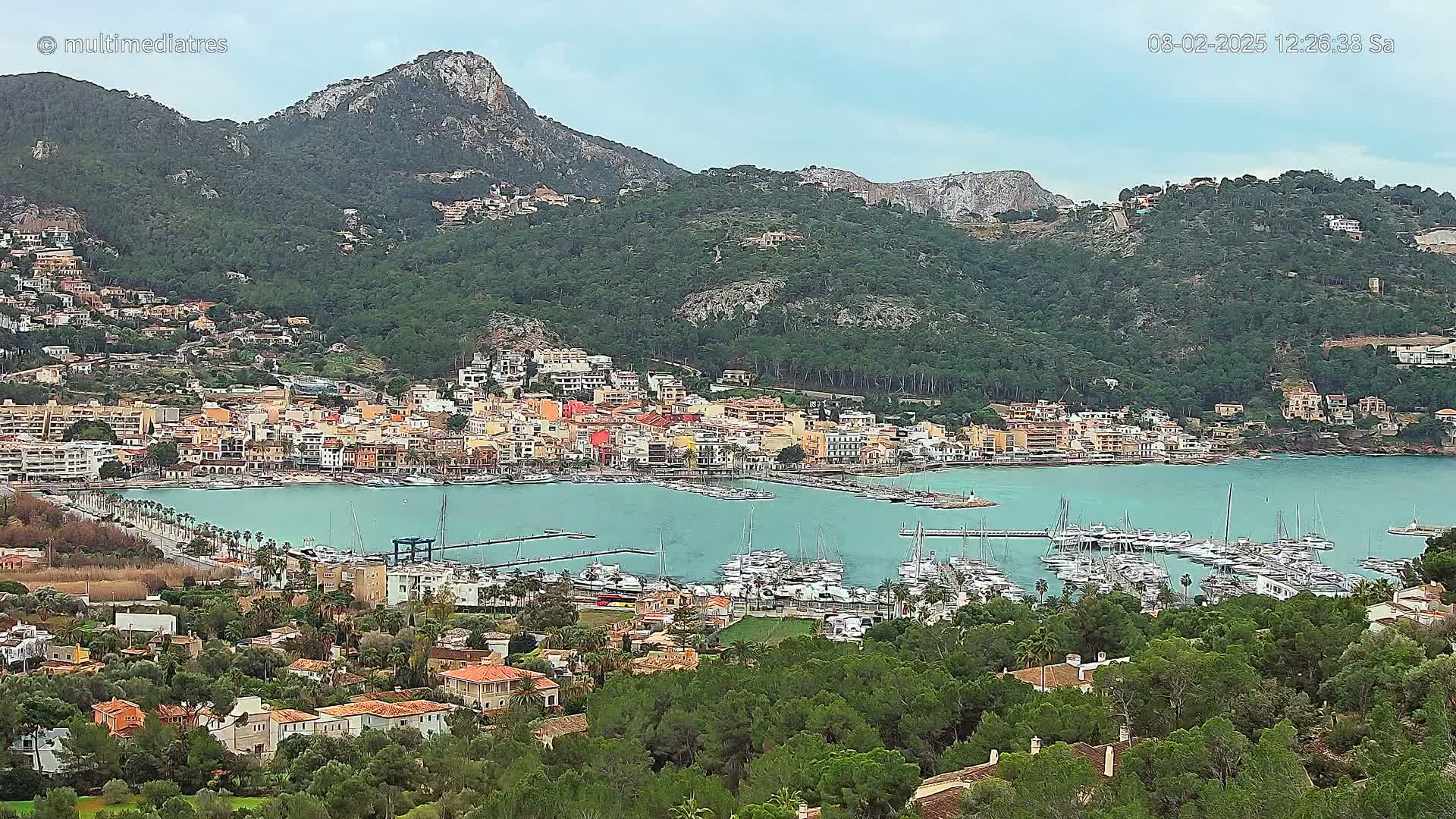 A picturesque coastal town nestled at the base of a verdant mountain range, under a mostly cloudy sky, features a bustling marina filled with numerous boats.