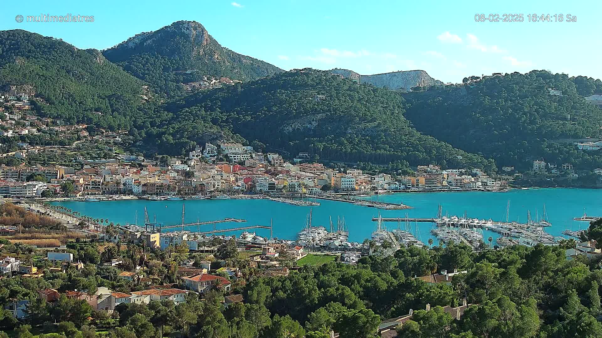 A picturesque coastal town nestled between green hills, with a vibrant blue harbor filled with numerous boats, under a clear, sunny sky.