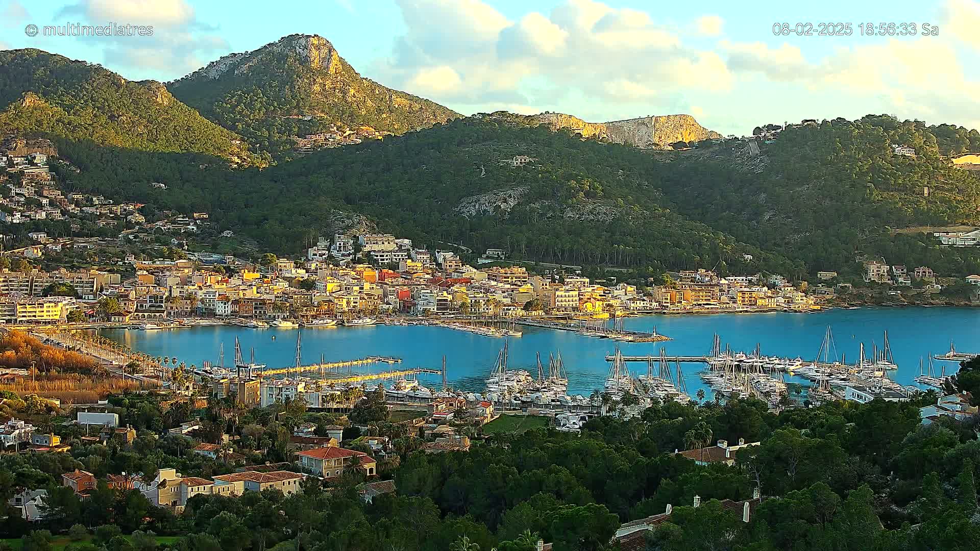 A picturesque coastal town nestled between mountains, with a harbor full of sailboats, under a partly cloudy sky.