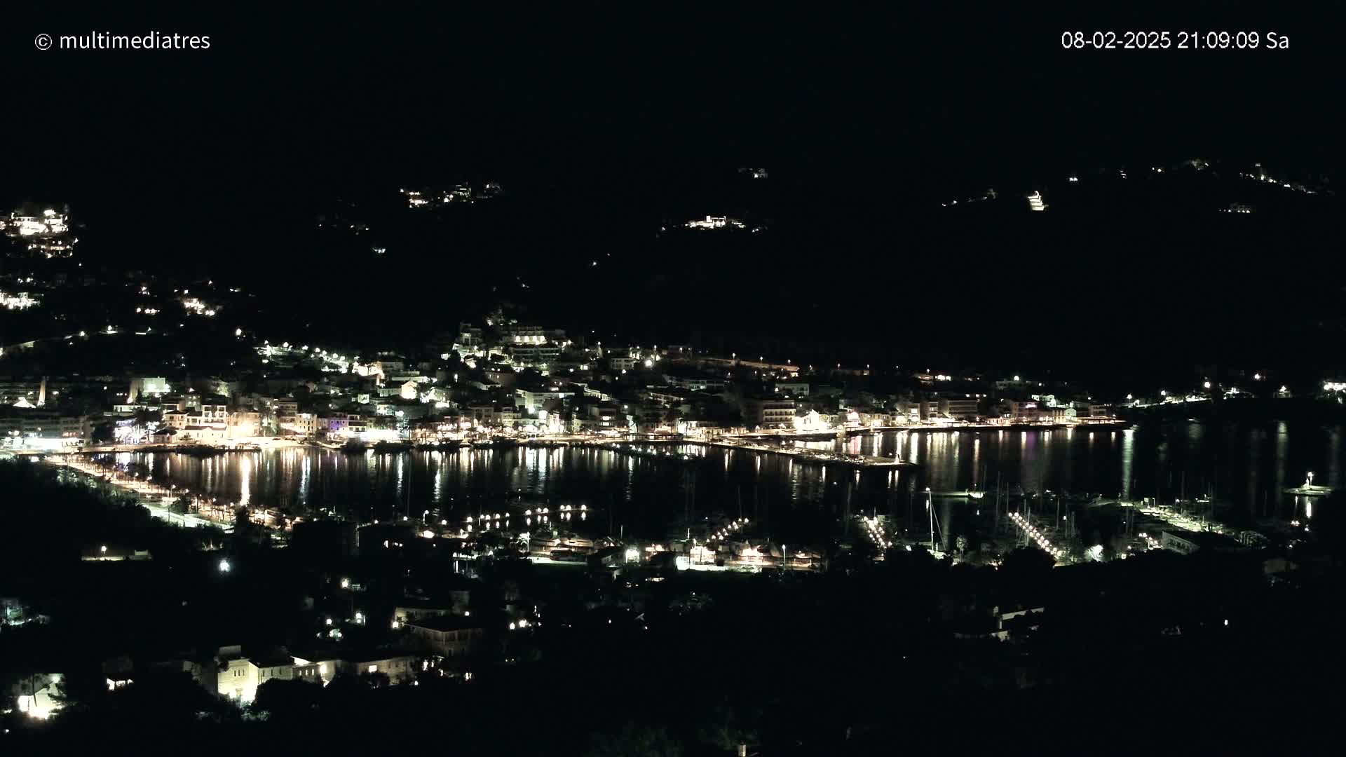 A nighttime view of a coastal town illuminated by lights, nestled along a dark body of water under a clear sky.