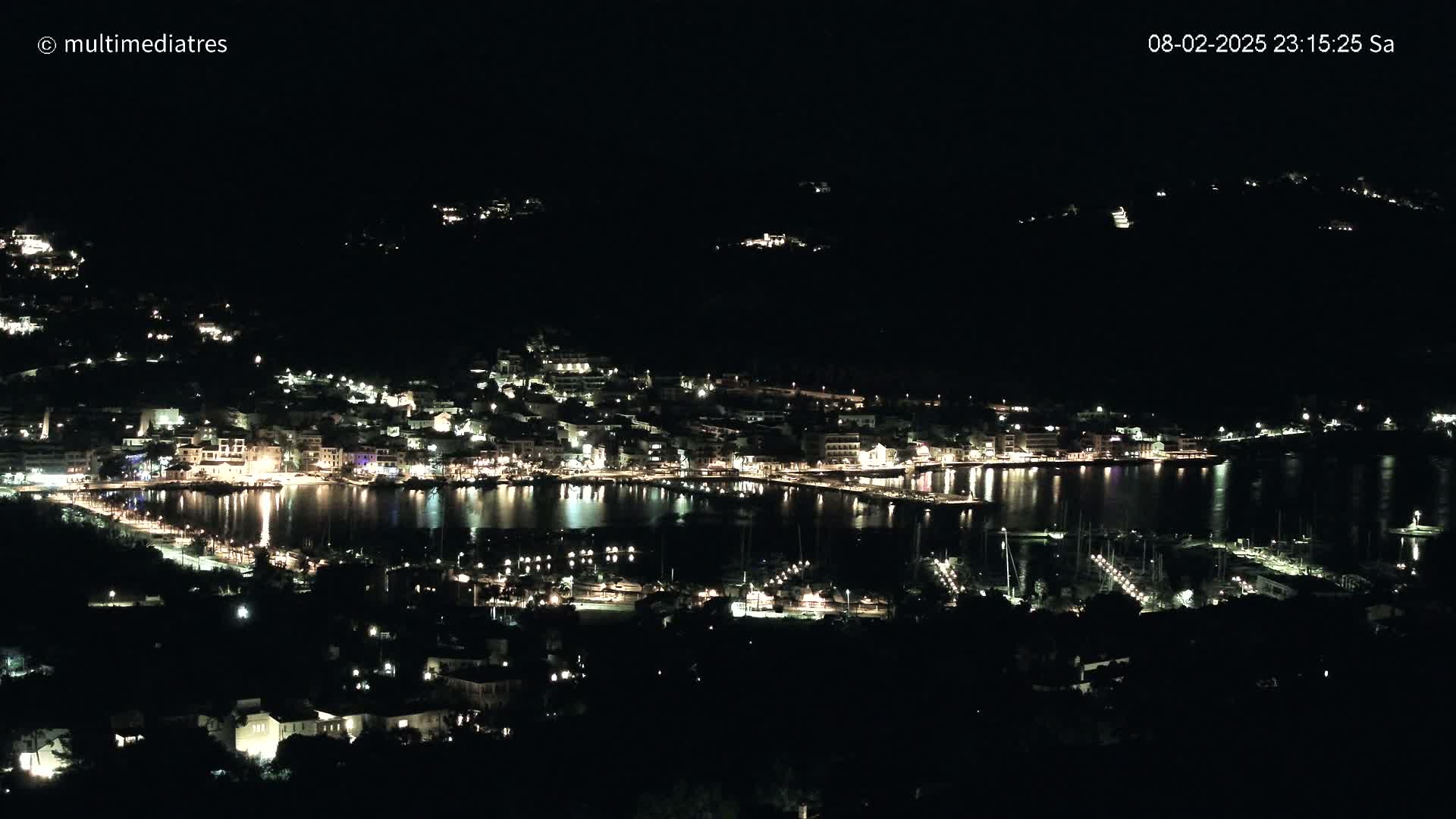 A nighttime, aerial view shows a coastal town with its lights illuminating the harbor and surrounding hills.