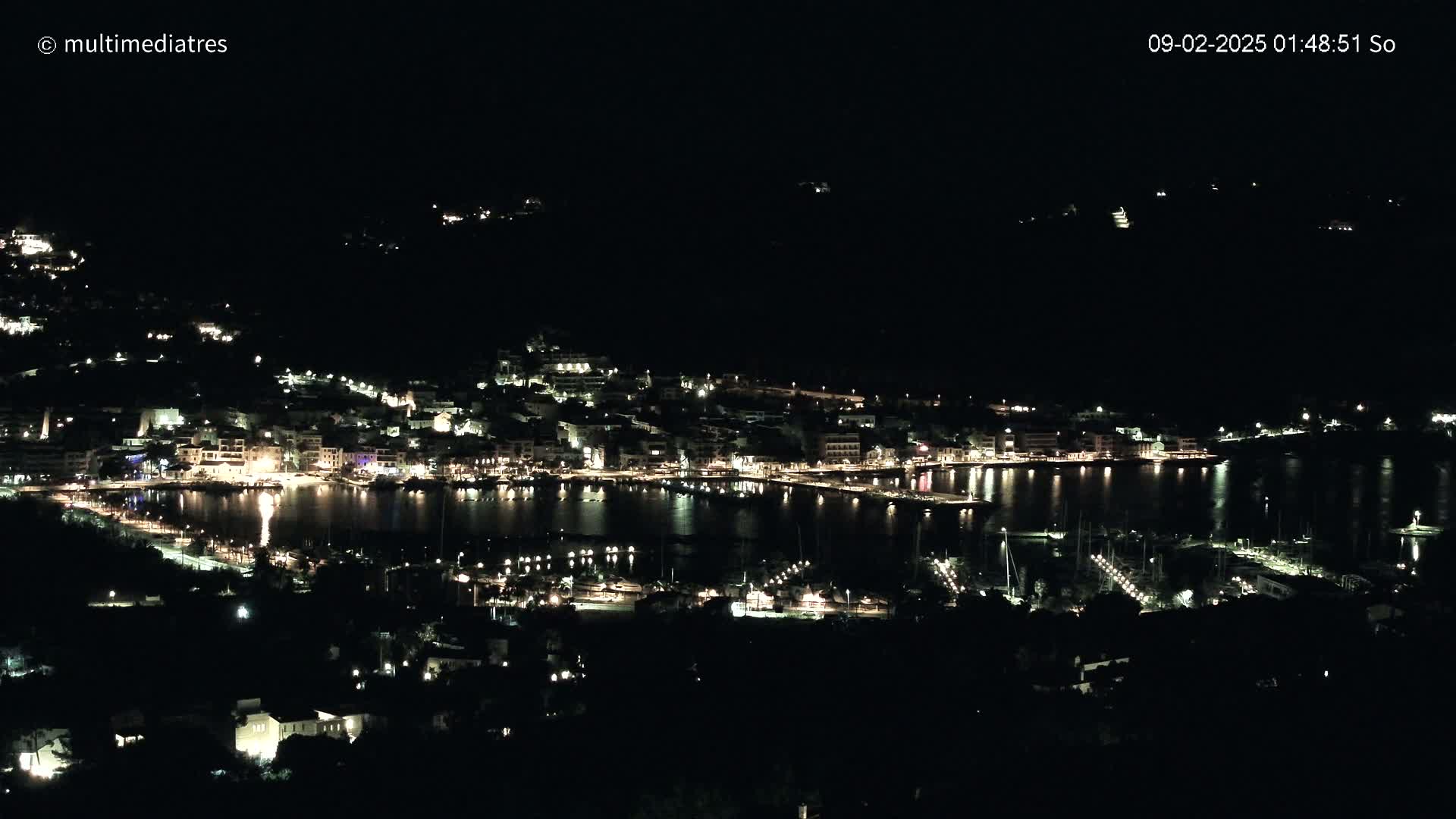 A nighttime view of a harbor town with many lights illuminating buildings and boats along a calm body of water.