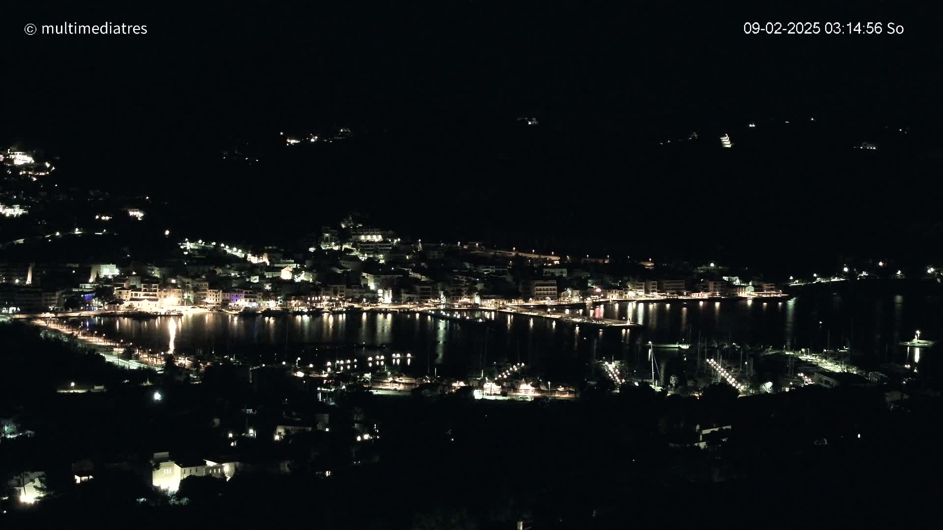 A nighttime view of a coastal town illuminated by lights, reflecting on a calm body of water.