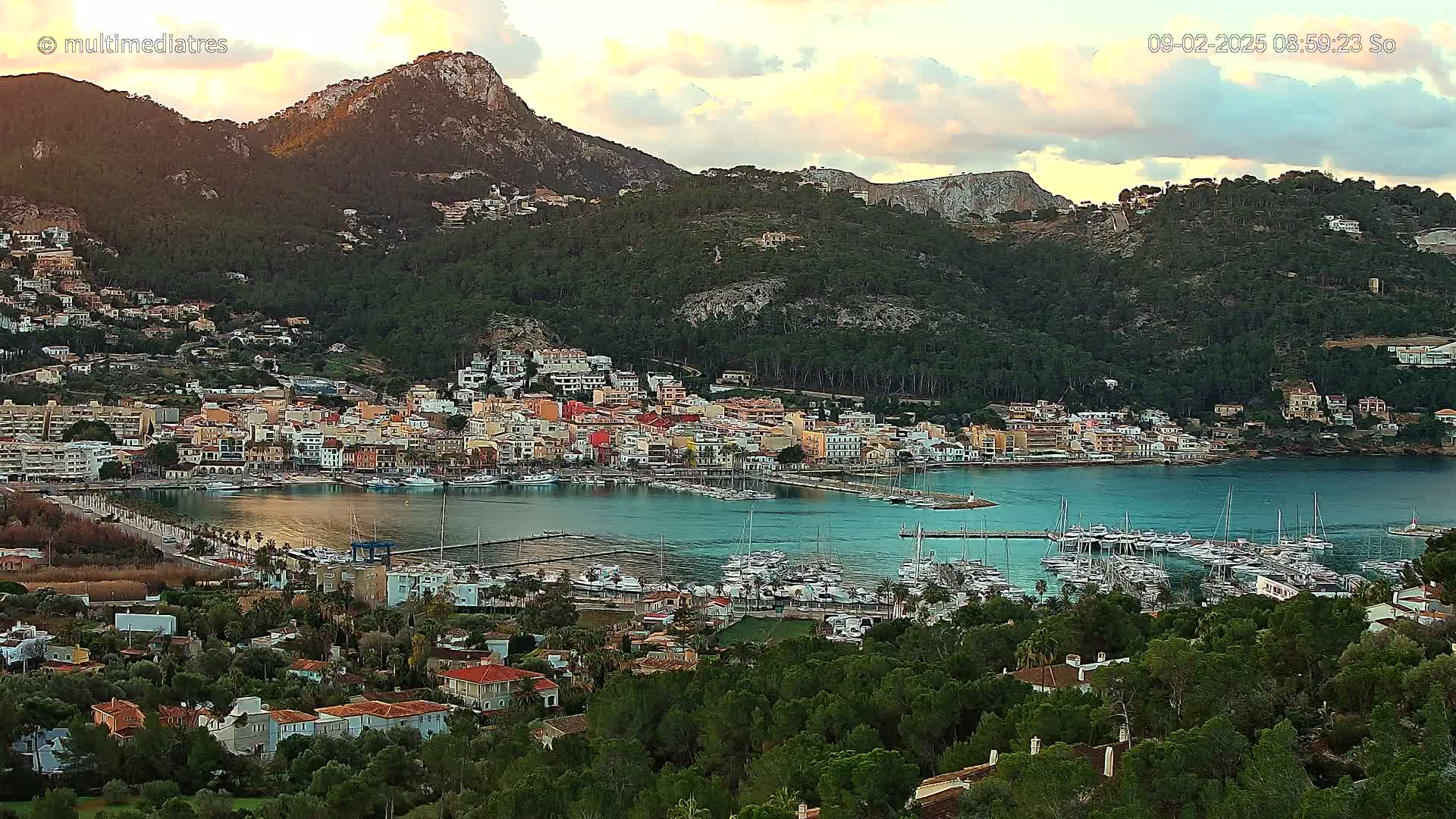 A coastal town with a marina filled with boats sits nestled at the base of a mountain range under a partly cloudy sunset sky.