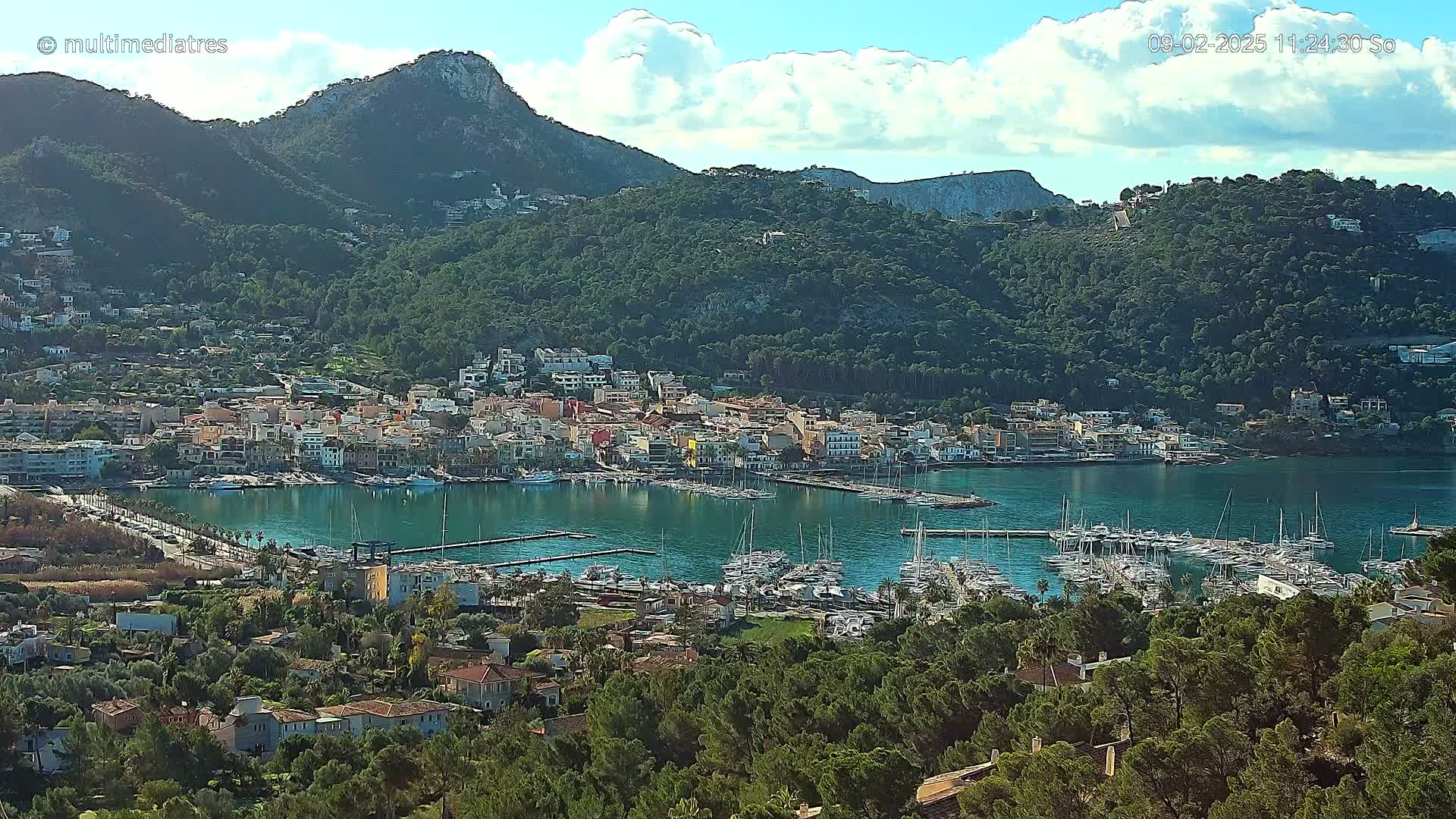 A coastal town nestled between verdant hills, with a harbor filled with sailboats under a partly cloudy sky.