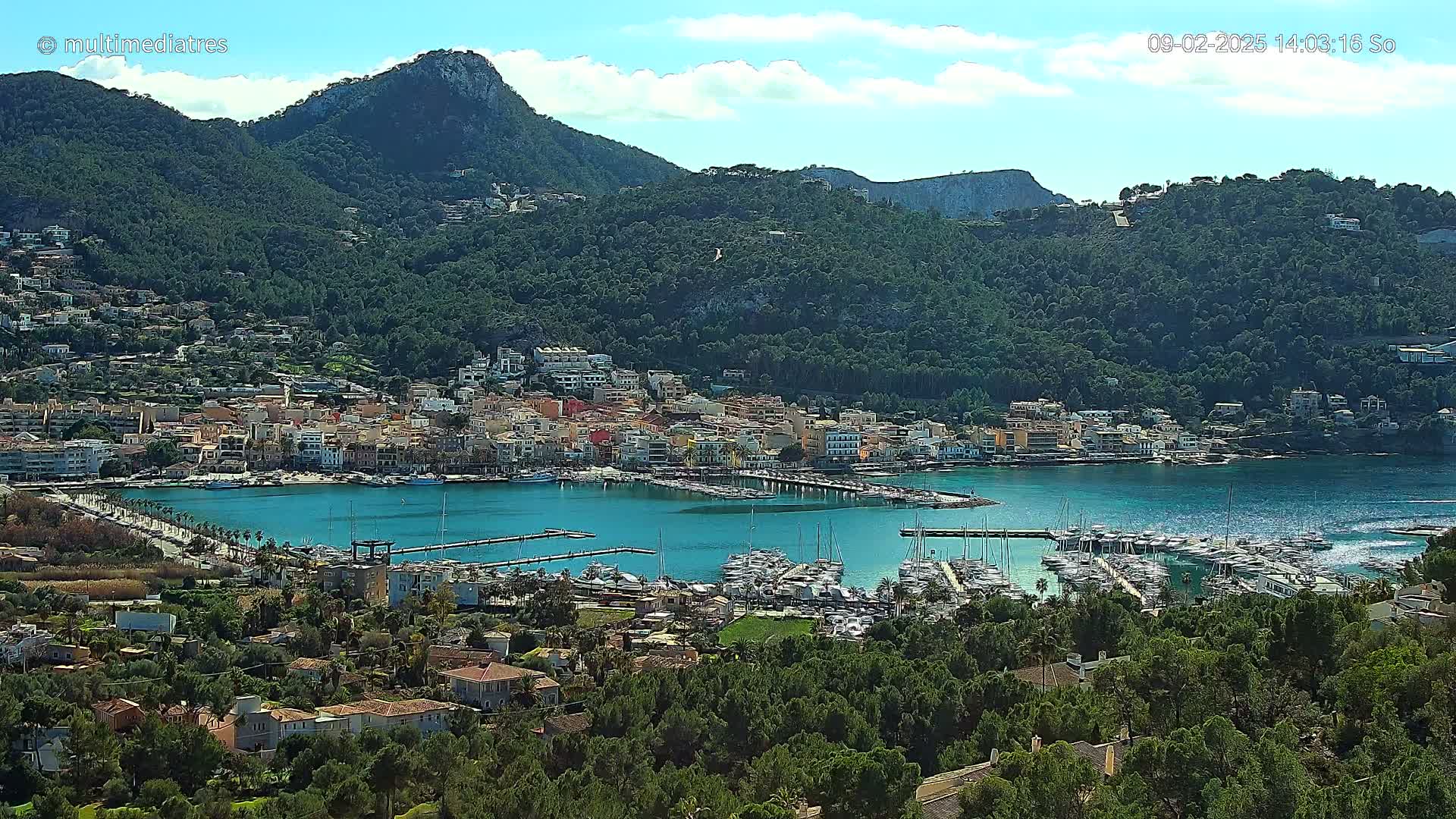 A picturesque coastal town nestled between verdant hills, featuring a calm harbor filled with numerous boats under a partly sunny sky.