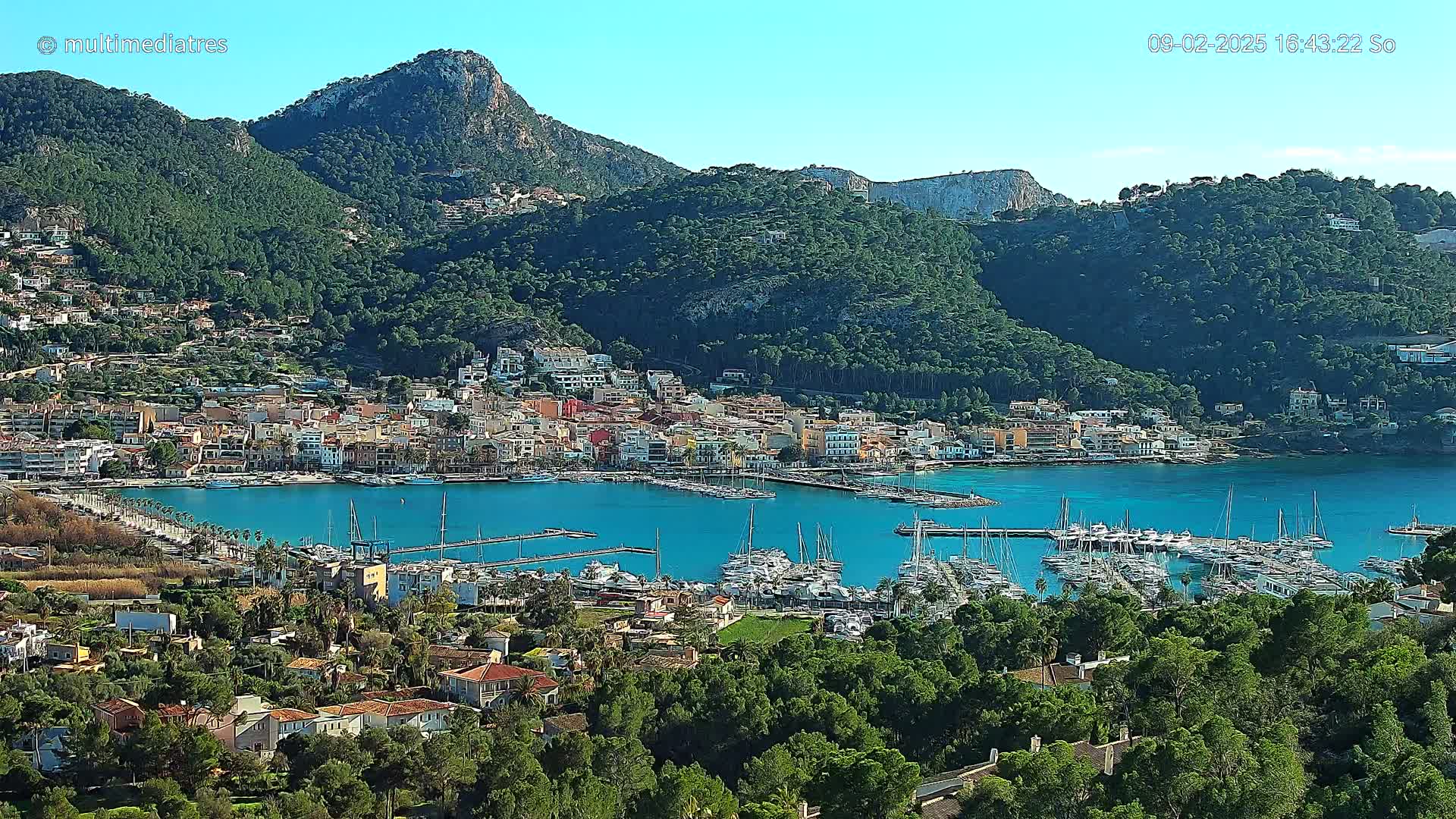 A picturesque coastal town nestled between lush green hills and a calm blue harbor filled with sailboats, under a clear sunny sky.