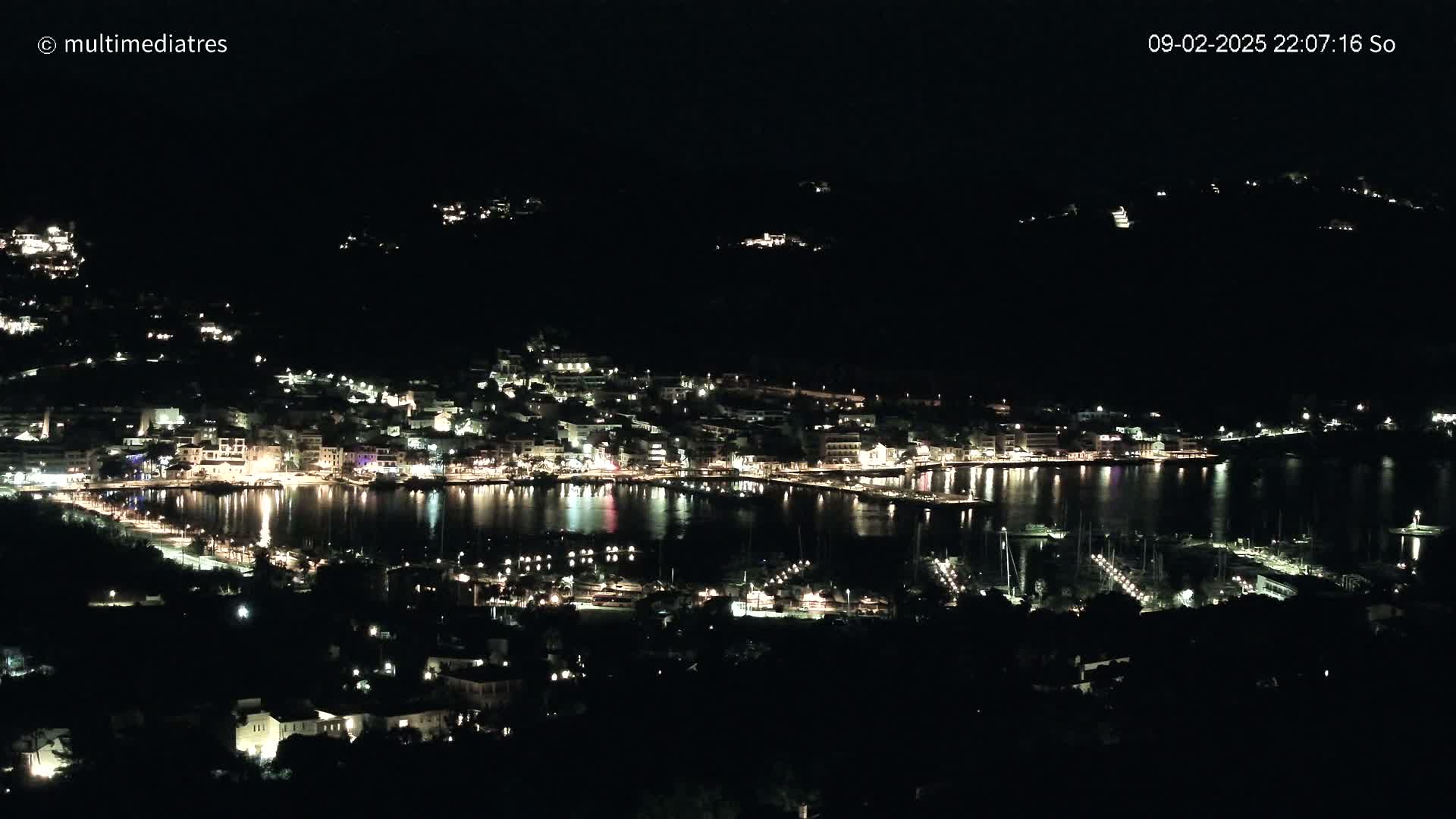 A nighttime, high-angle view shows a coastal town illuminated with many lights, nestled along a calm body of water dotted with boats.