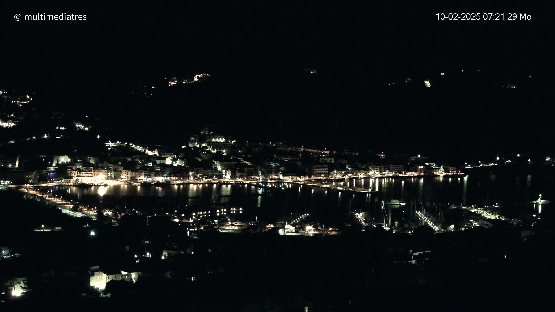 A nighttime view of a coastal town with its lights illuminating the harbor and surrounding buildings, under a clear, dark sky.