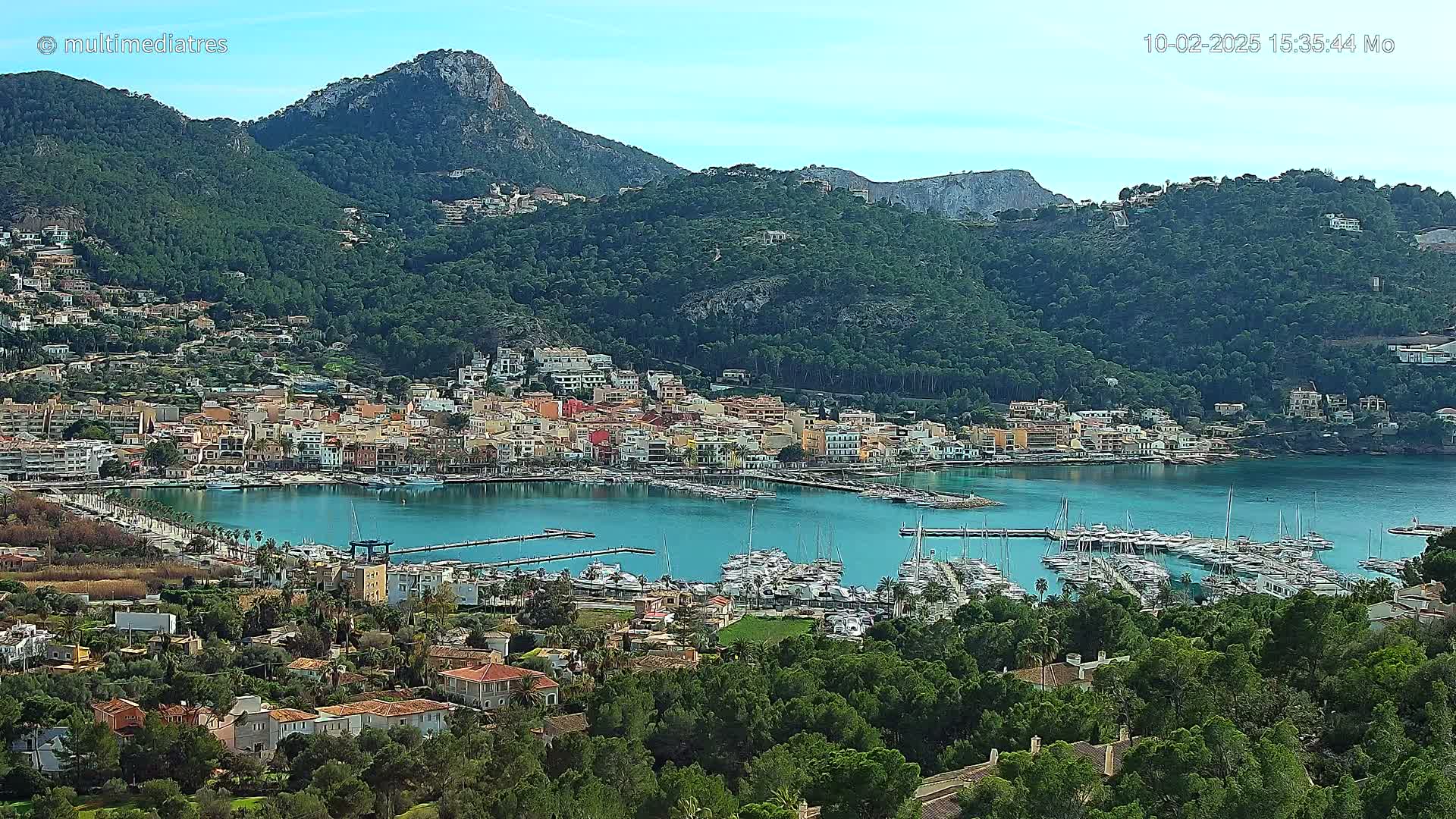 A picturesque coastal town nestled in a green, mountainous region, with a calm blue harbor filled with numerous yachts and boats under a clear sky.